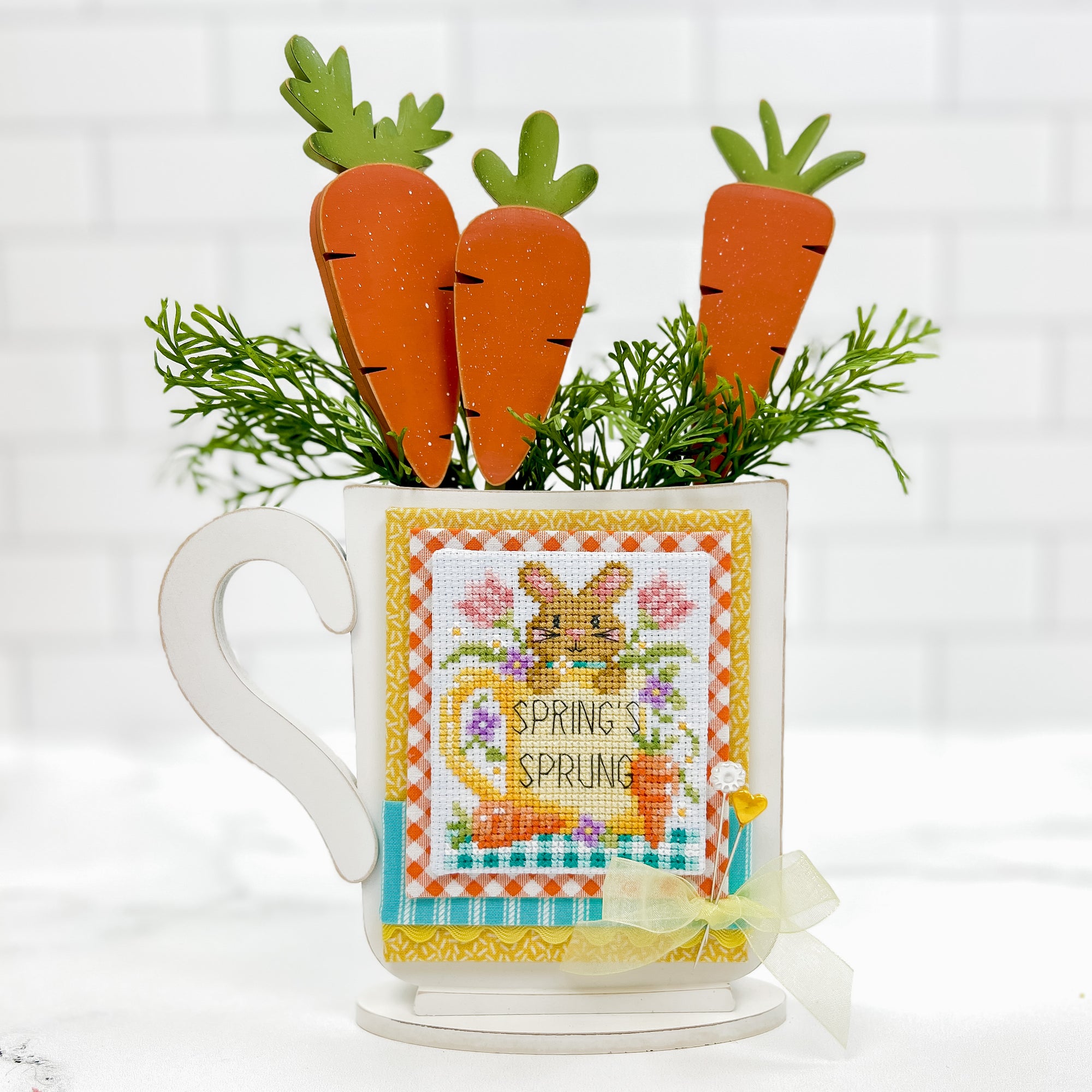 Cross stitch display mug with orange wood carrots inside the mug. Cross stitch design is a bunny by Shannon Christine Designs