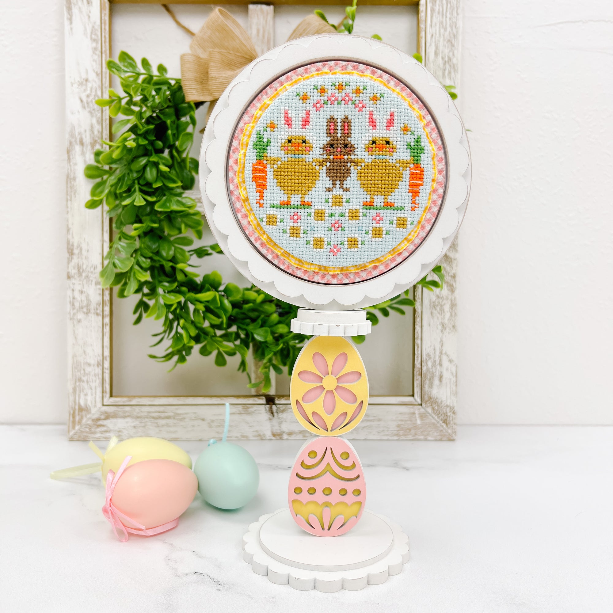 Round cross stitch display on an egg shaped pedestal with a finished Easter cross stitch by Stitching with the Housewives.