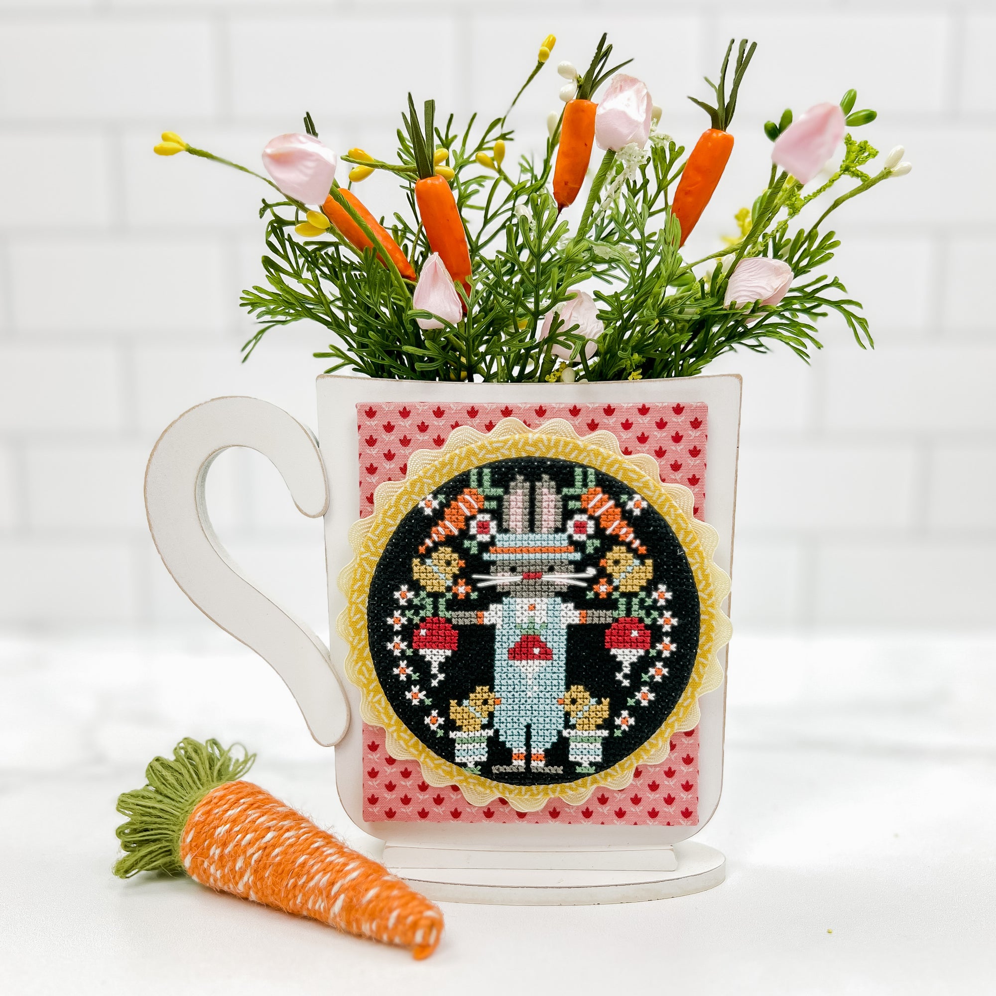 Mug cross stitch display with greenery in the top. Cross stitch is a bunny and chicks by Stitching with the Housewives
