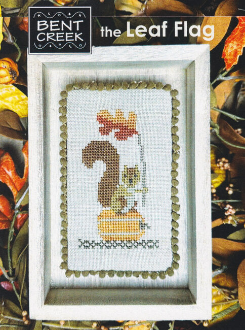 Cross-stitched design of a squirrel with a  oak leaf sitting on a fall pumpkin