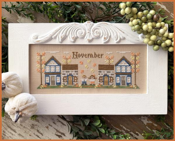 Cross-stitched scene with houses and 'November' text depicting an Autumnal scene of Fall foliage 