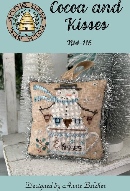 Cross-stitched snowman pillow with a cocoa banner in a cocoa mug.