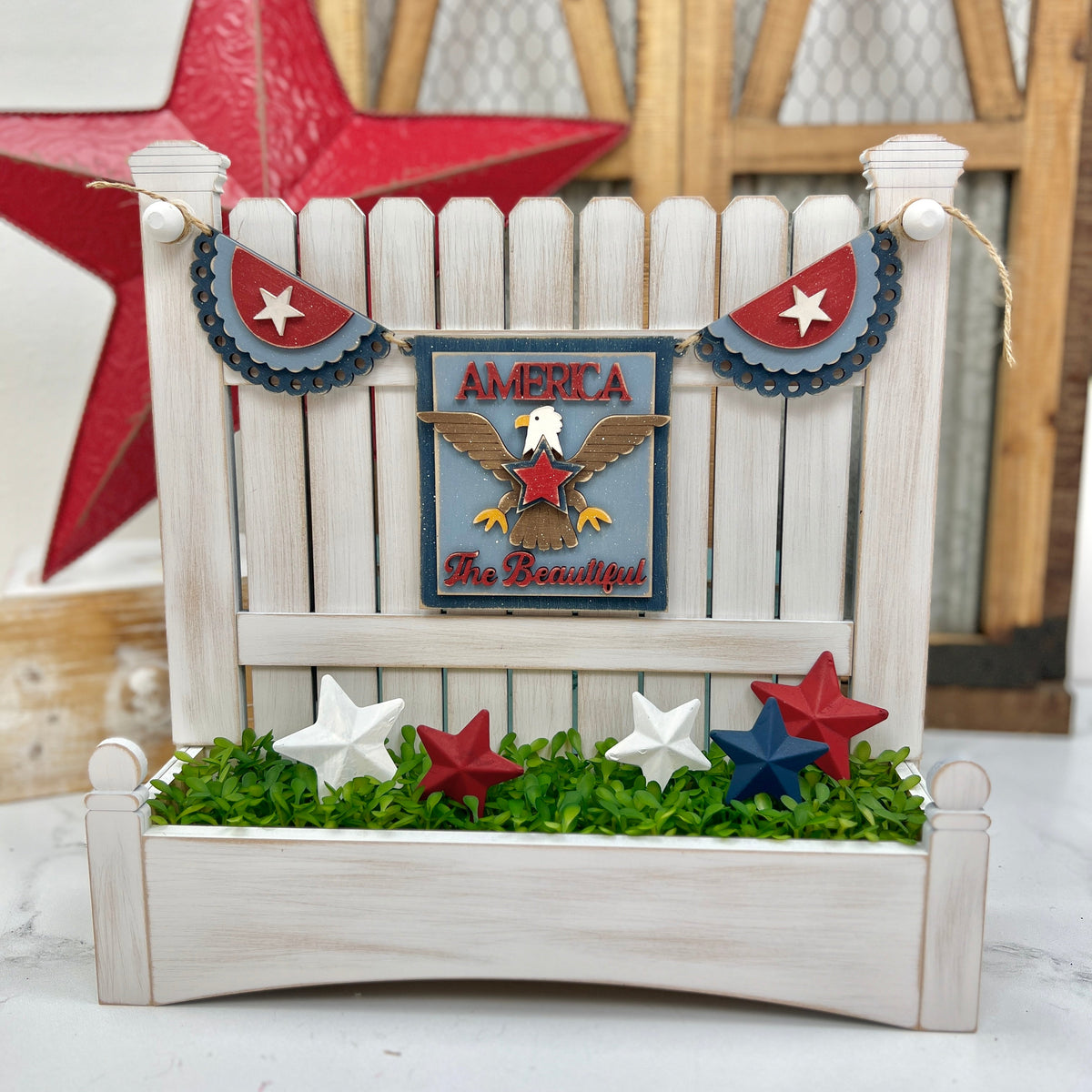 Wooden picket fence display decorated with a DIY patriotic garland featuring scalloped red, white, and blue bunting, an eagle plaque that says “America the Beautiful,” and painted wooden stars in red, white, and blue nestled in faux greenery.
