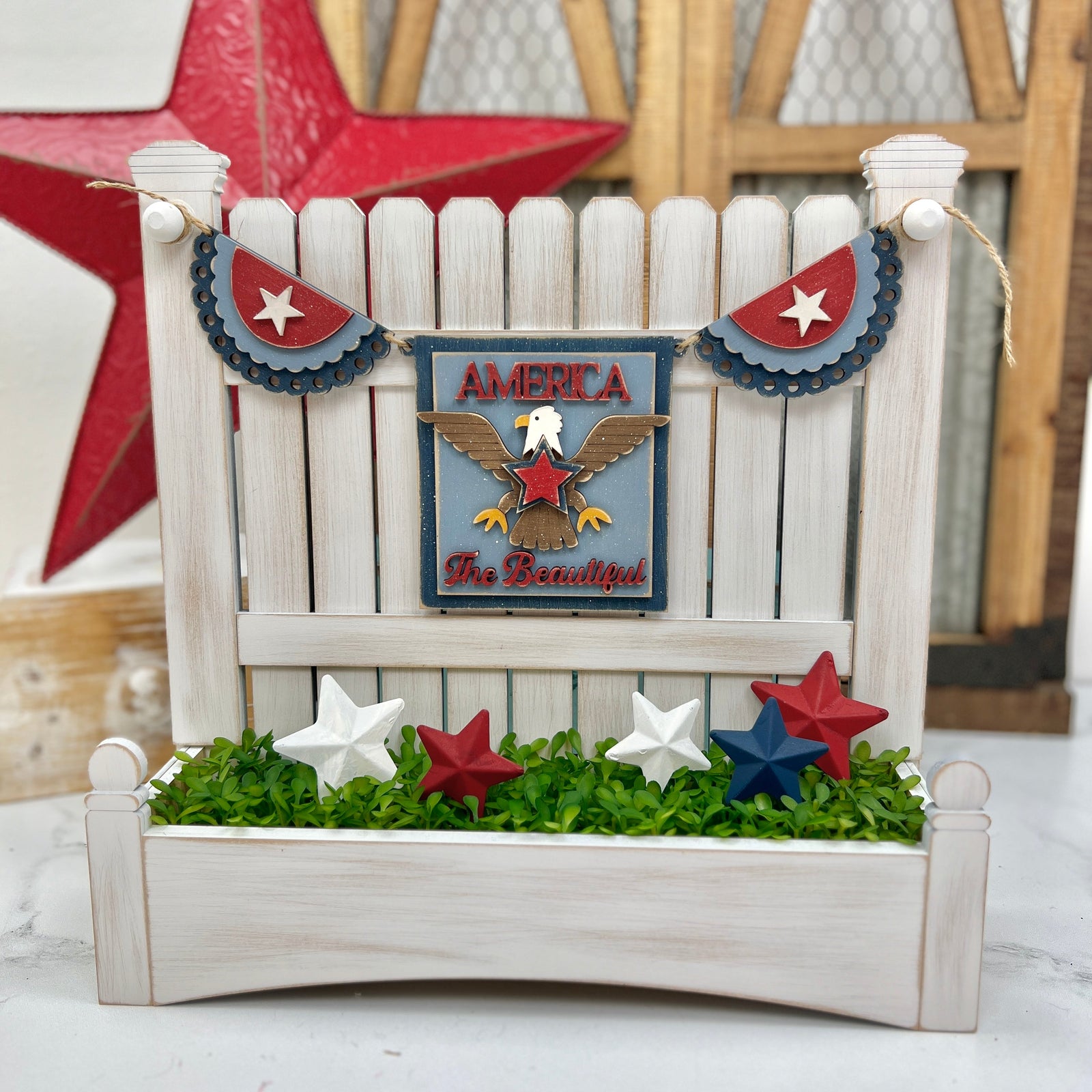 Wooden picket fence display decorated with a DIY patriotic garland featuring scalloped red, white, and blue bunting, an eagle plaque that says “America the Beautiful,” and painted wooden stars in red, white, and blue nestled in faux greenery.