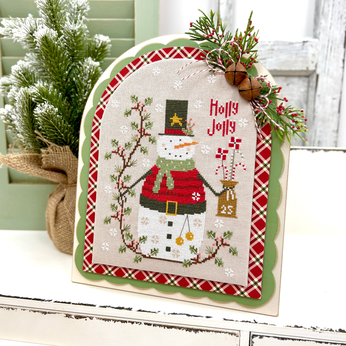Arbor cross stitch display in pre-painted sage green with a scalloped arch border, styled with a finished Holly Jolly snowman cross stitch and red plaid fabric.