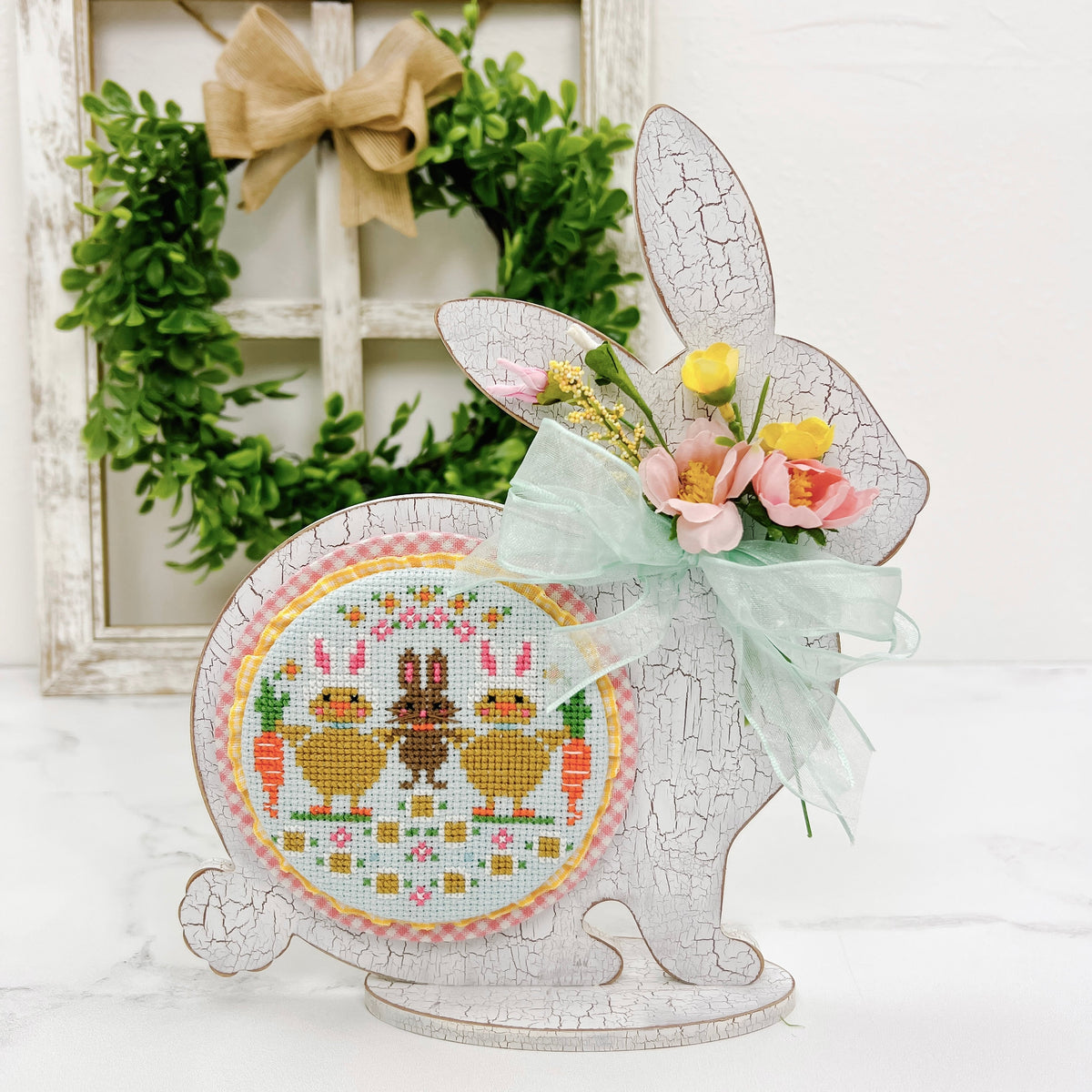 Sitting chunky bunny wood cross stitch display backer with a finished Easter cross stitch by Stitching with the Housewives.