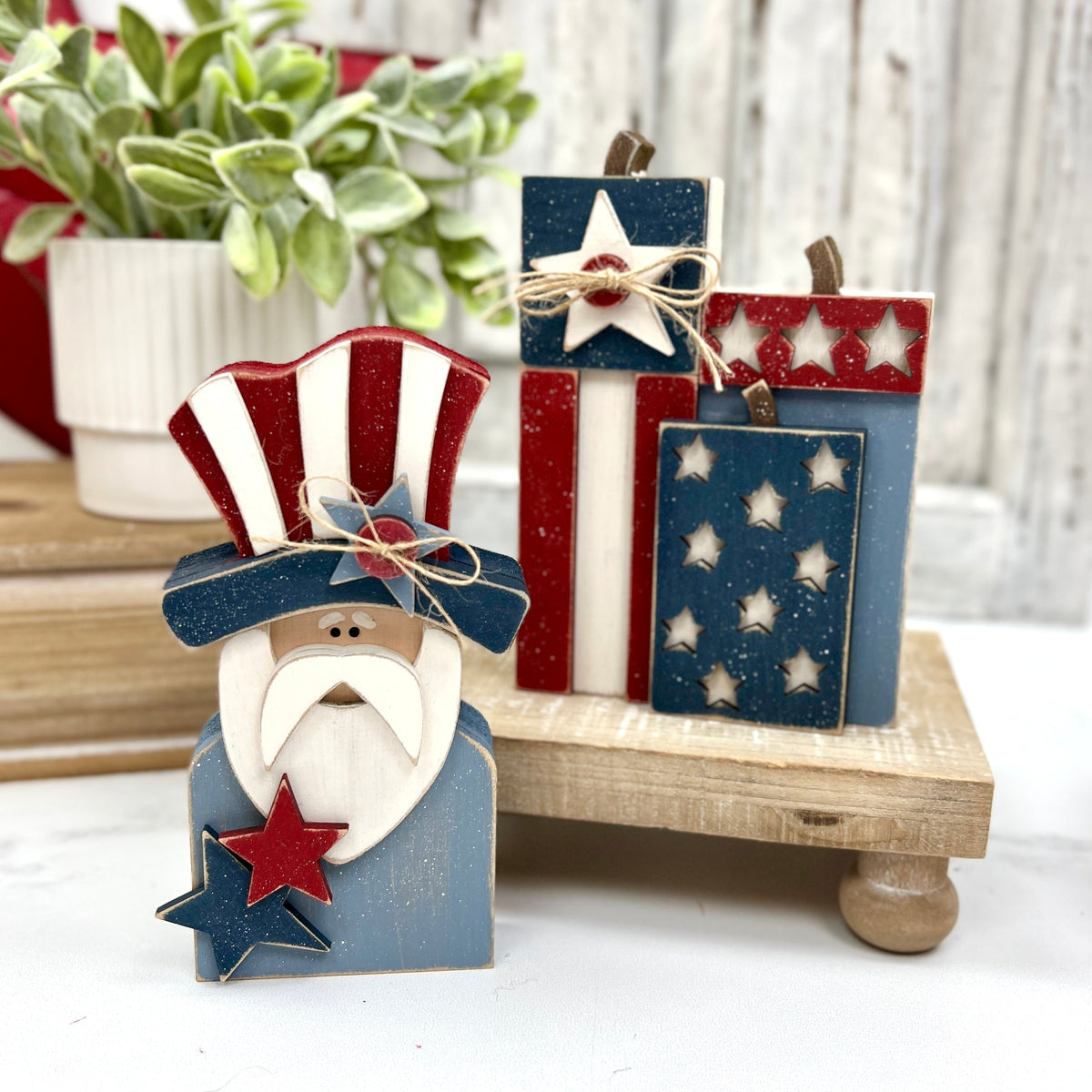 Chunky wooden Uncle Sam figure with a red, white, and blue hat and matching star accents, displayed next to three stacked wooden “fireworks” painted in patriotic colors with star cutouts and twine bows, set on a small wooden riser.