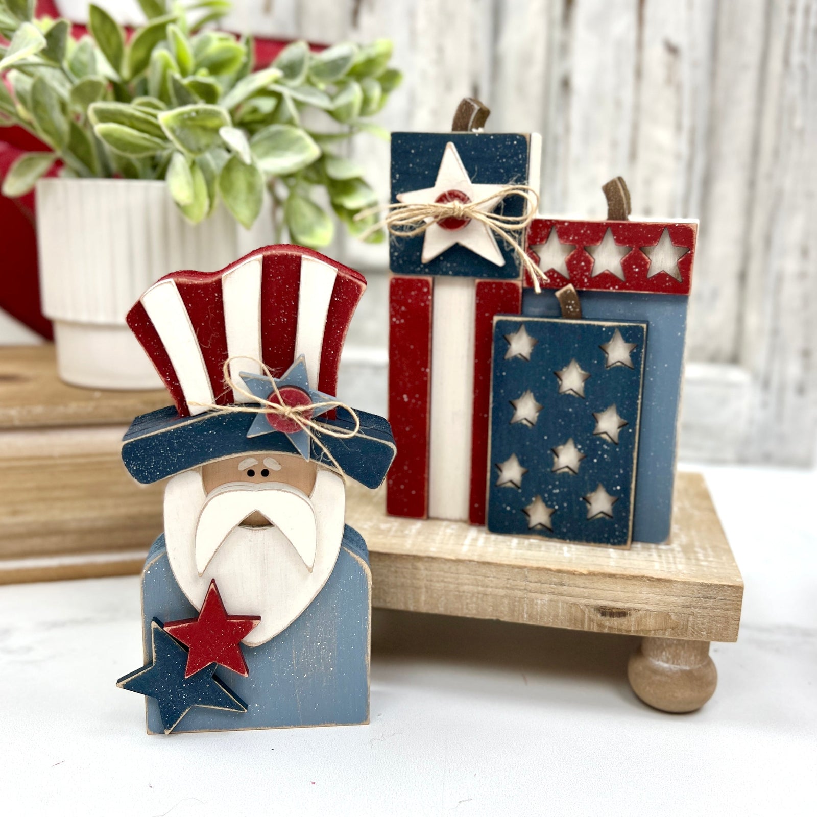 Chunky wooden Uncle Sam figure with a red, white, and blue hat and matching star accents, displayed next to three stacked wooden “fireworks” painted in patriotic colors with star cutouts and twine bows, set on a small wooden riser.