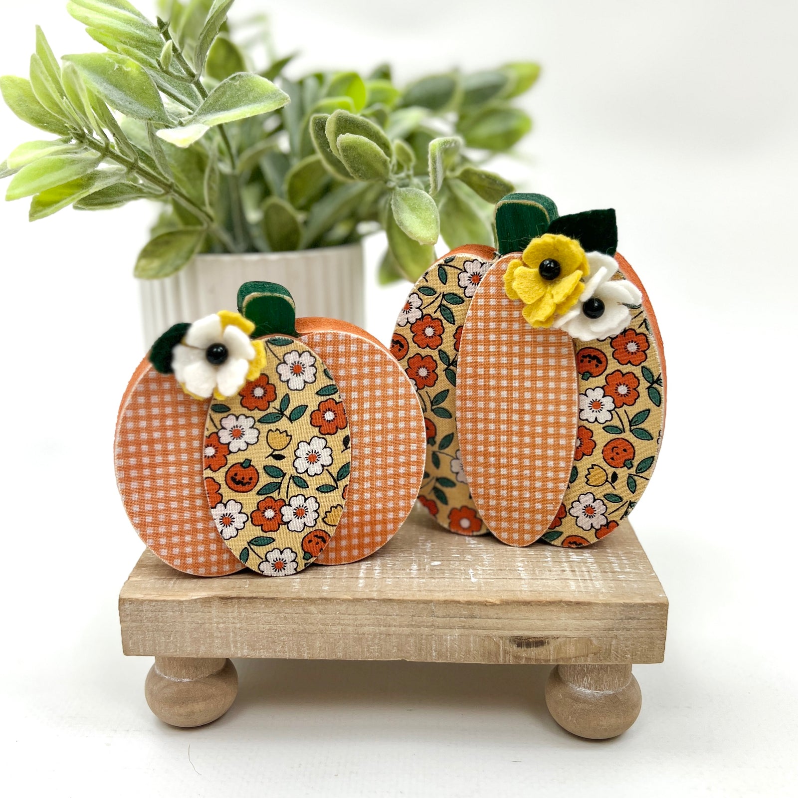 Decorative pumpkins with floral patterns on a wooden stand with a plant in the background.
