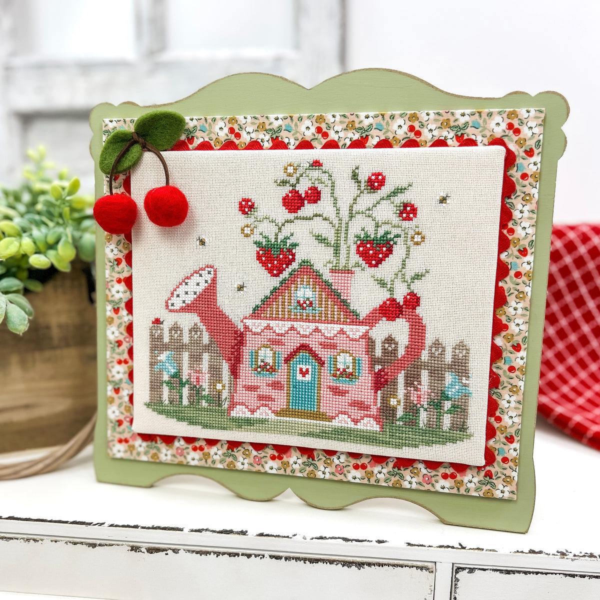 Finished Cottage in the Berry Patch cross stitch design mounted on a pre-painted sage green scalloped wood display, featuring a pink cottage, strawberry plants, and a felt cherry embellishment.