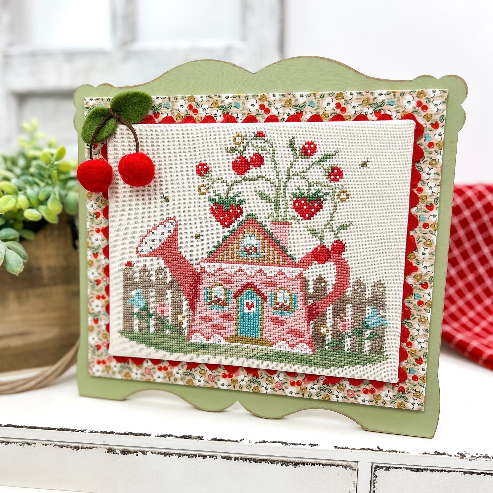 Finished Cottage in the Berry Patch cross stitch design mounted on a pre-painted sage green scalloped wood display, featuring a pink cottage, strawberry plants, and a felt cherry embellishment.