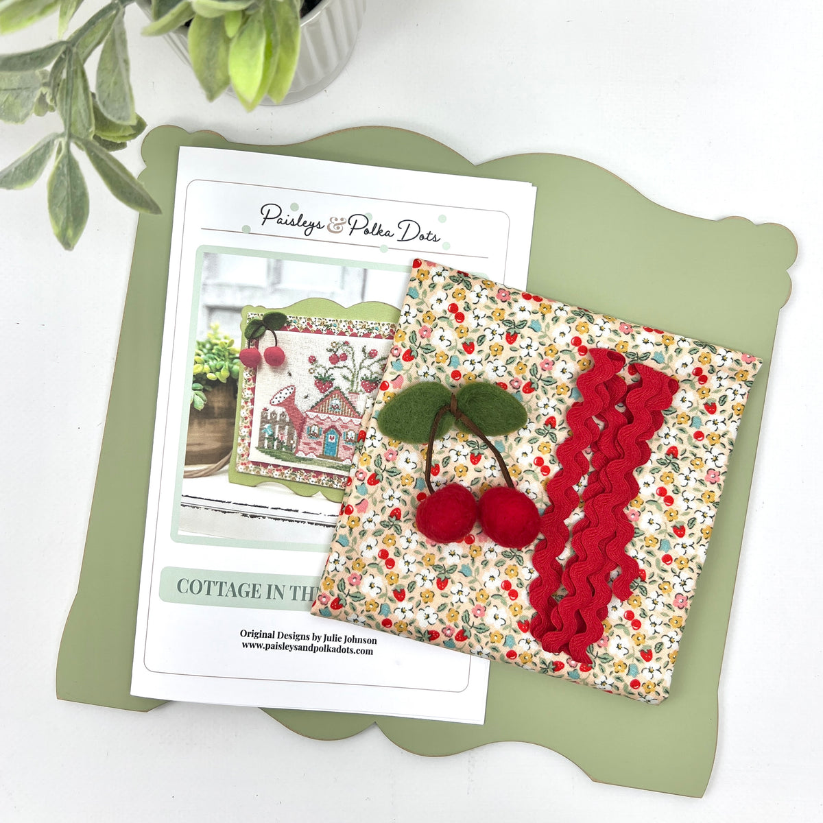 Cottage in the Berry Patch cross stitch display kit by Paisleys &amp; Polka Dots, showing the pattern cover, cherry-and-strawberry print fabric, red rickrack, and felt cherries on a sage green scalloped wood display.