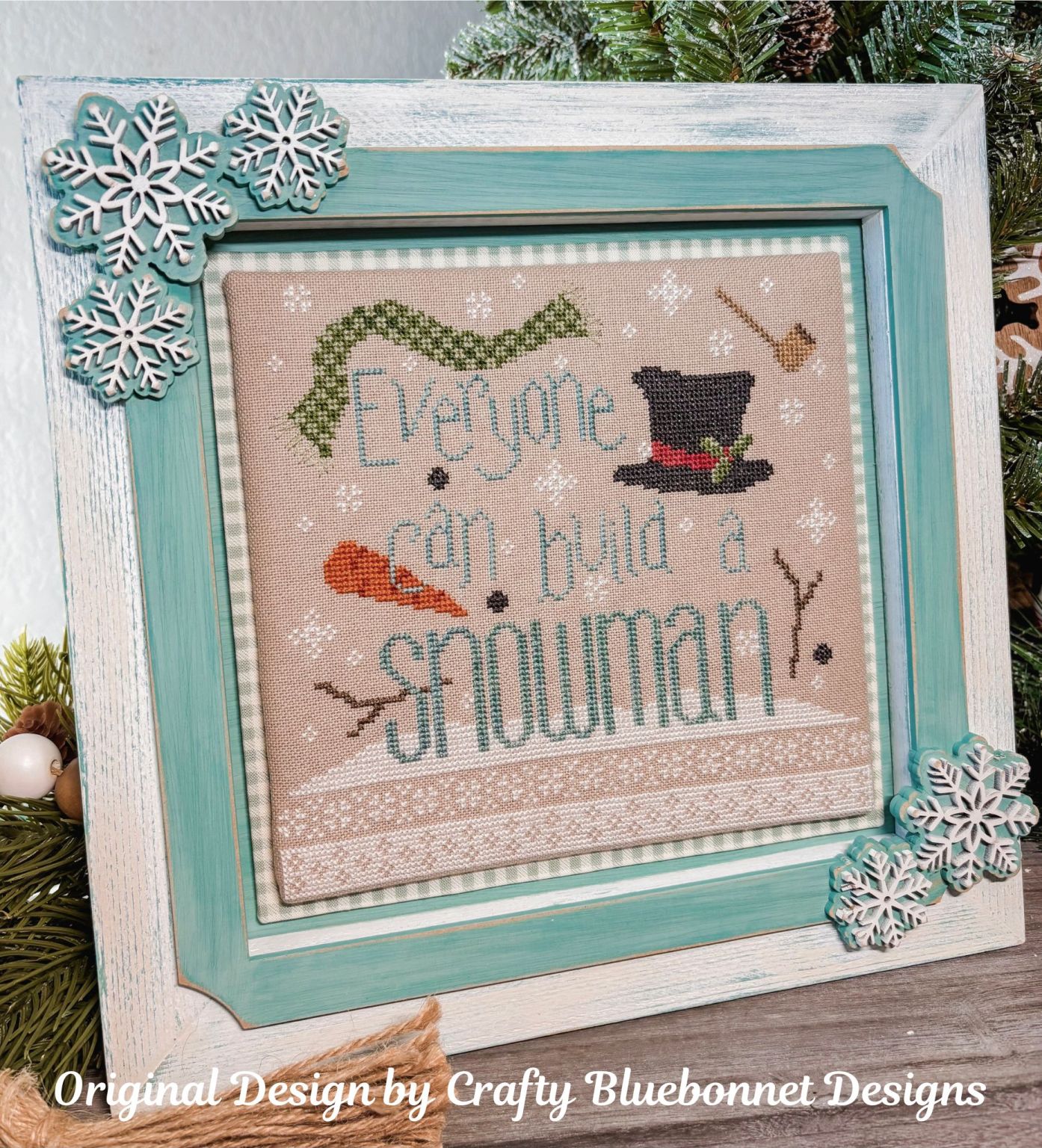 Decorative cross-stitch piece with 'Everyone can build a snowman' text, framed with a winter themed snowflake display frame.
