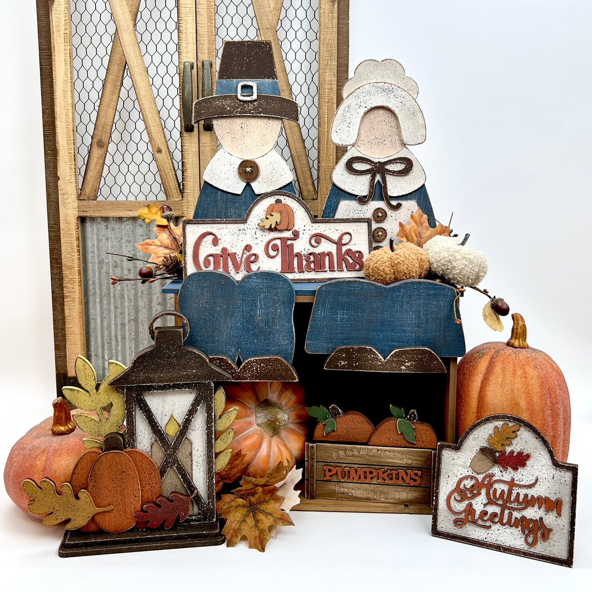 Rustic fall display featuring primitive pilgrim shelf sitters, a lantern with pumpkin and leaf accents, and wooden signs reading “Give Thanks,” “Pumpkins,” and “Autumn Greetings,” surrounded by faux pumpkins and autumn leaves.