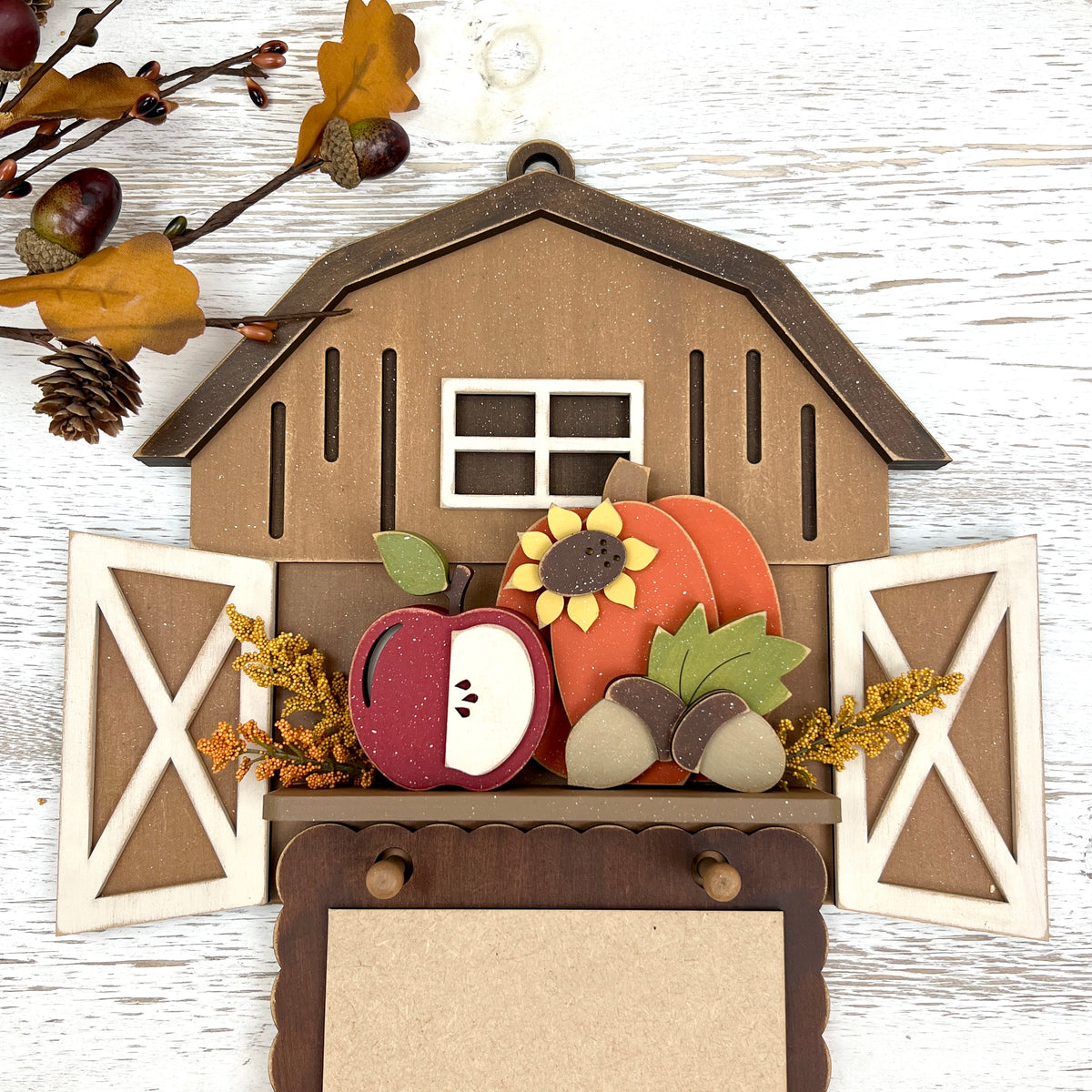 Autumn-themed wooden wall hanging shaped like a barn with open doors, featuring layered fall decorations including a red apple, orange pumpkin, sunflower, acorns, and leaves, displayed on a white wood background.