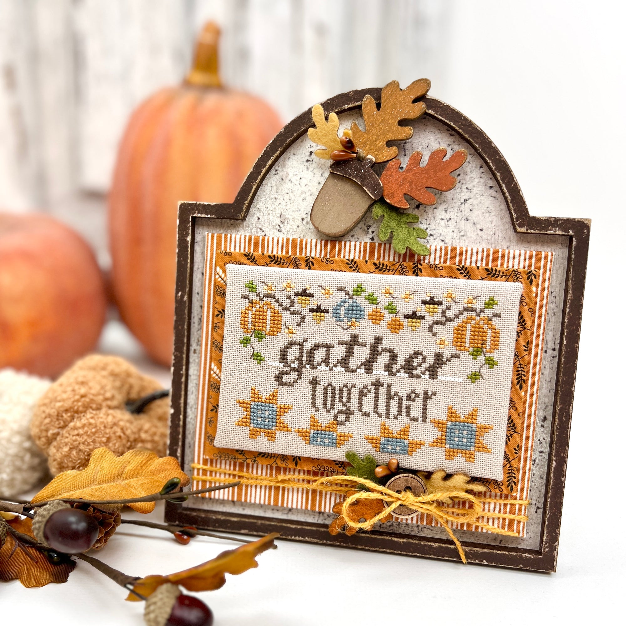 Unfinished wood cross stitch display with an arched top, acorn and leaf embellishment, and “Gather Together” cross stitched piece mounted in the center. DIY frame with easel stand, painted in rustic fall colors.