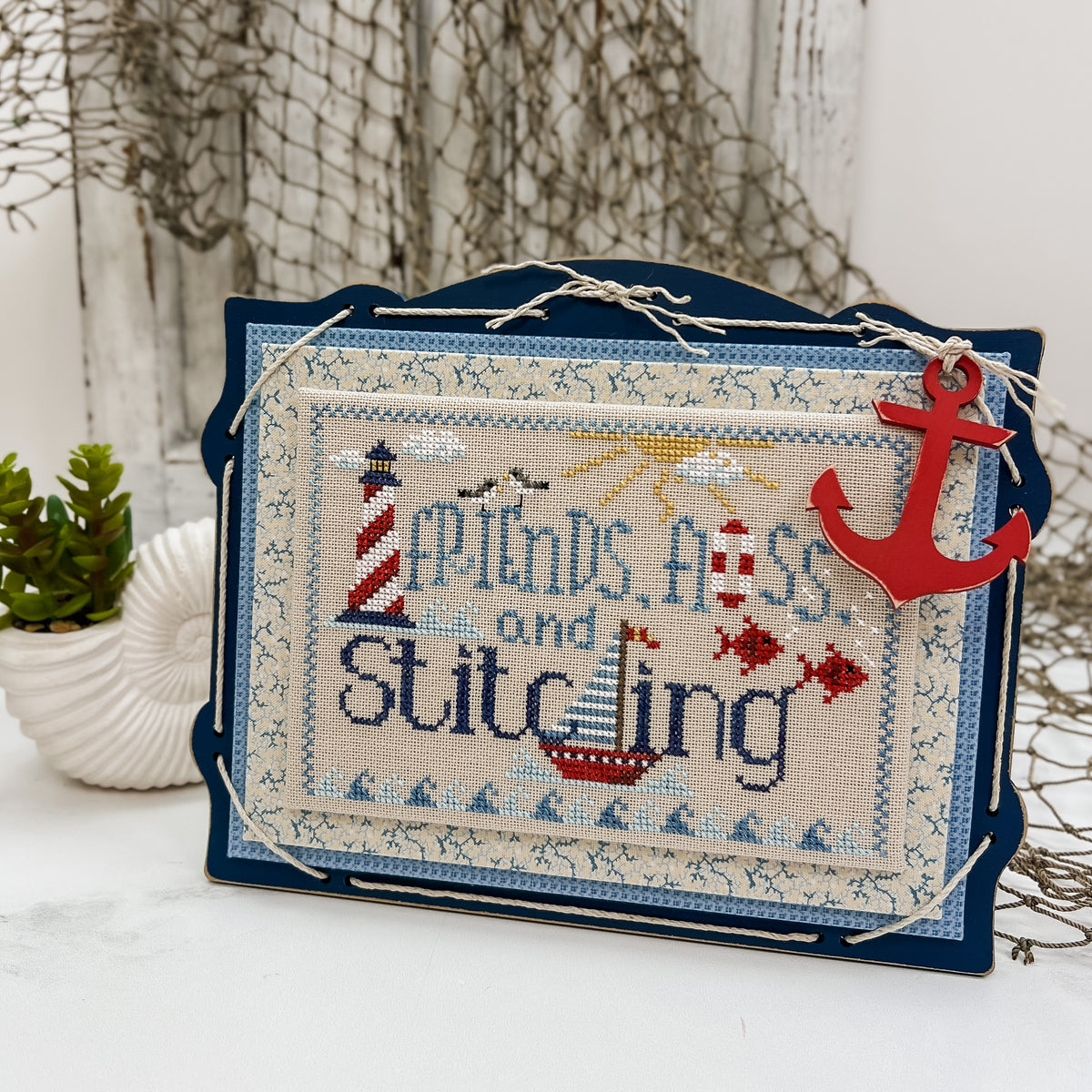 Nautical themed Friends, Floss, and Stitching cross stitch project displayed on a dark blue pre-painted board with blue finishing fabrics, twine lacing, and a red anchor charm.