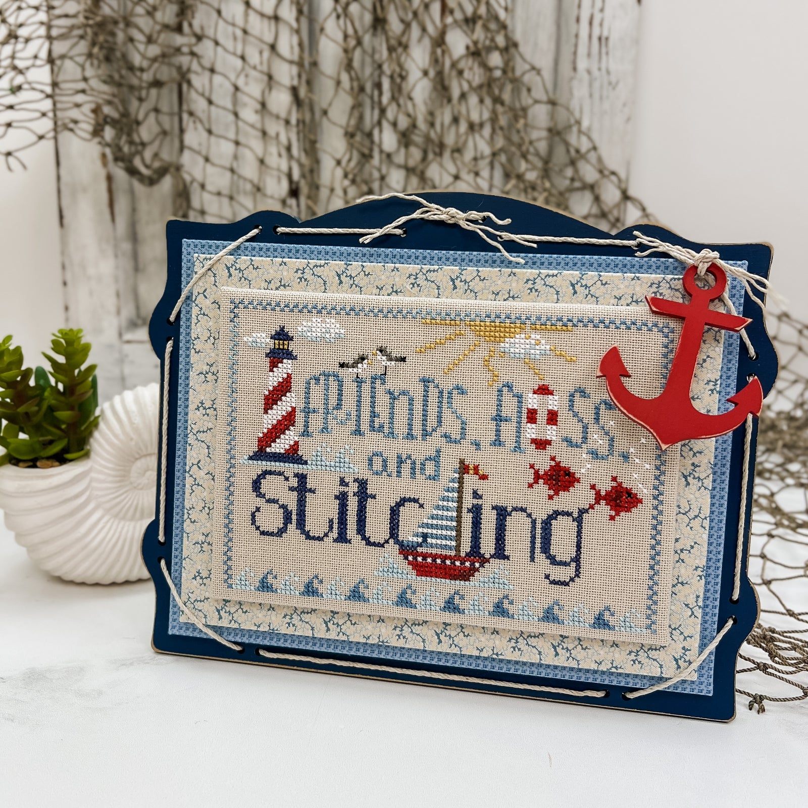 Nautical themed Friends, Floss, and Stitching cross stitch project displayed on a dark blue pre-painted board with blue finishing fabrics, twine lacing, and a red anchor charm.