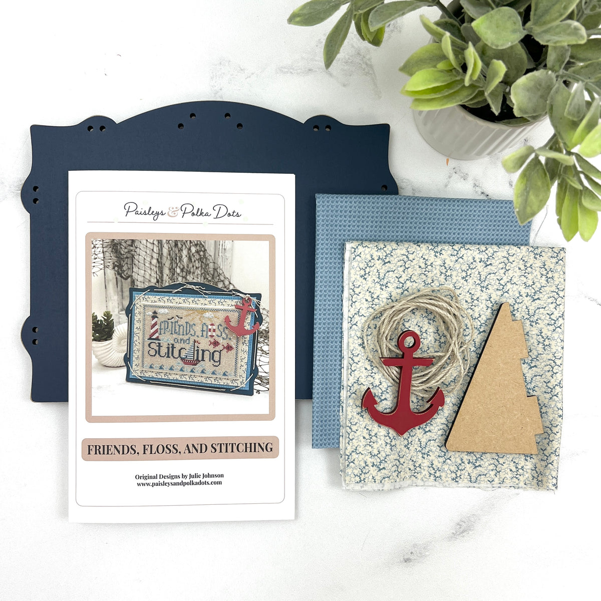 Nautical themed Friends, Floss, and Stitching cross stitch project displayed on a dark blue pre-painted board with blue finishing fabrics, twine lacing, and a red anchor charm.