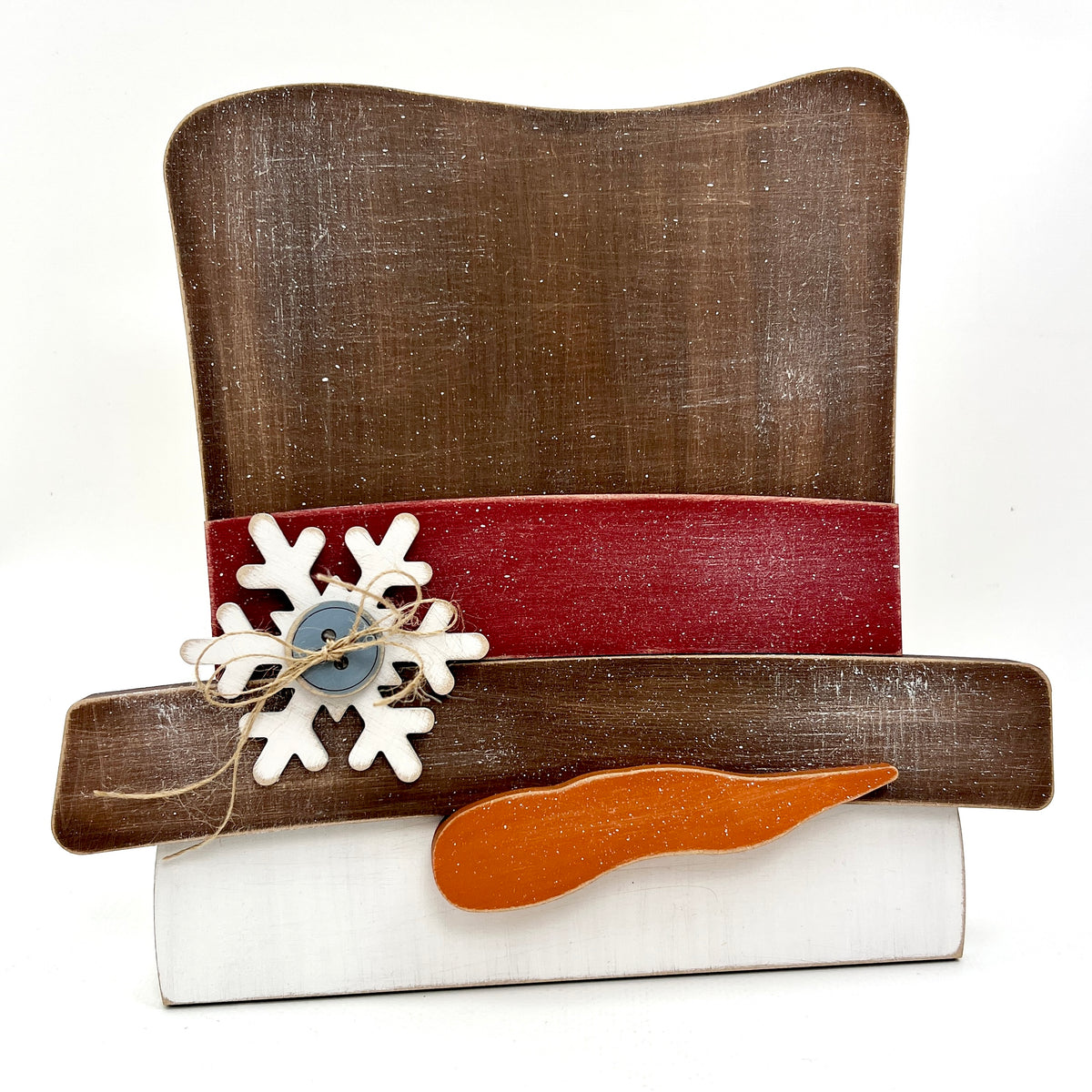 Decorative wood cross stitch display with a brown top hat, red band, white snowflake, blue button, and wooden paddle on a white background