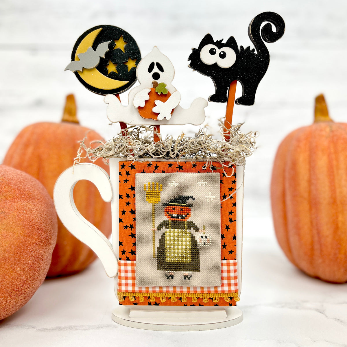 Halloween Wood mug decoration with a ghost. black cat, and moon with stars placed into the mug. Mug displays a finished cross stitch piece with a witch by Brenda Gervais. 