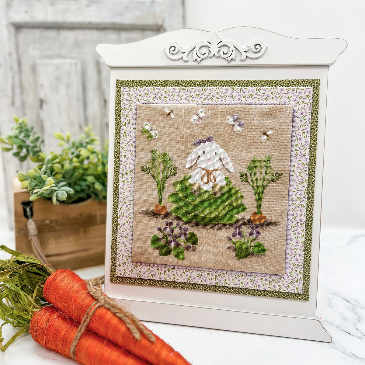 Pre-painted heirloom embroidery and cross stitch display frame with a neutral finish, decorative scroll top, layered fabric accents, and a bunny cross stitch displayed in the center.