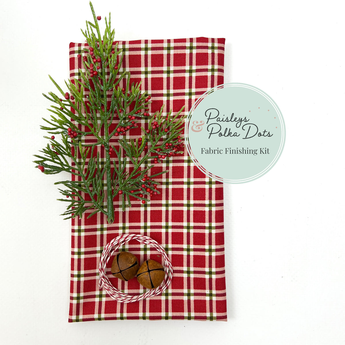 Paisleys &amp; Polka Dots fabric finishing kit for cross stitch, featuring red, green, and white plaid Christmas fabric styled with a pine sprig, red berries, rusty jingle bells, and red-and-white twine.
