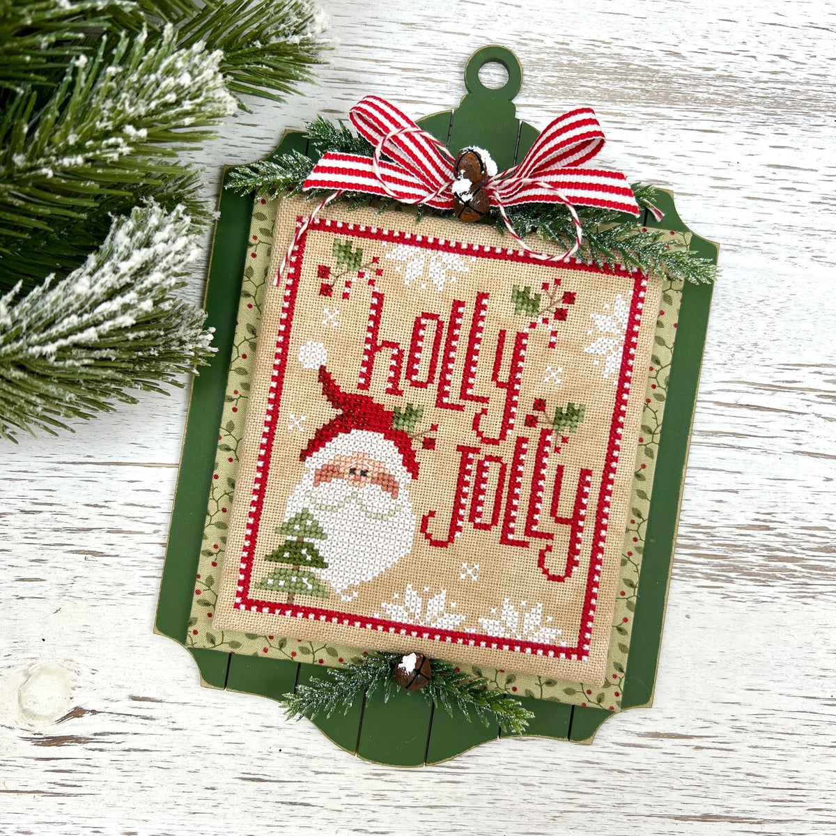 A Christmas cross-stitch ornament featuring a Santa Claus design and the words 'holly jolly' in red embroidery on a beige fabric background, mounted on a green wooden plaque with a red-and-white striped ribbon bow at the top, photographed on a white wood surface with pine branches around it.