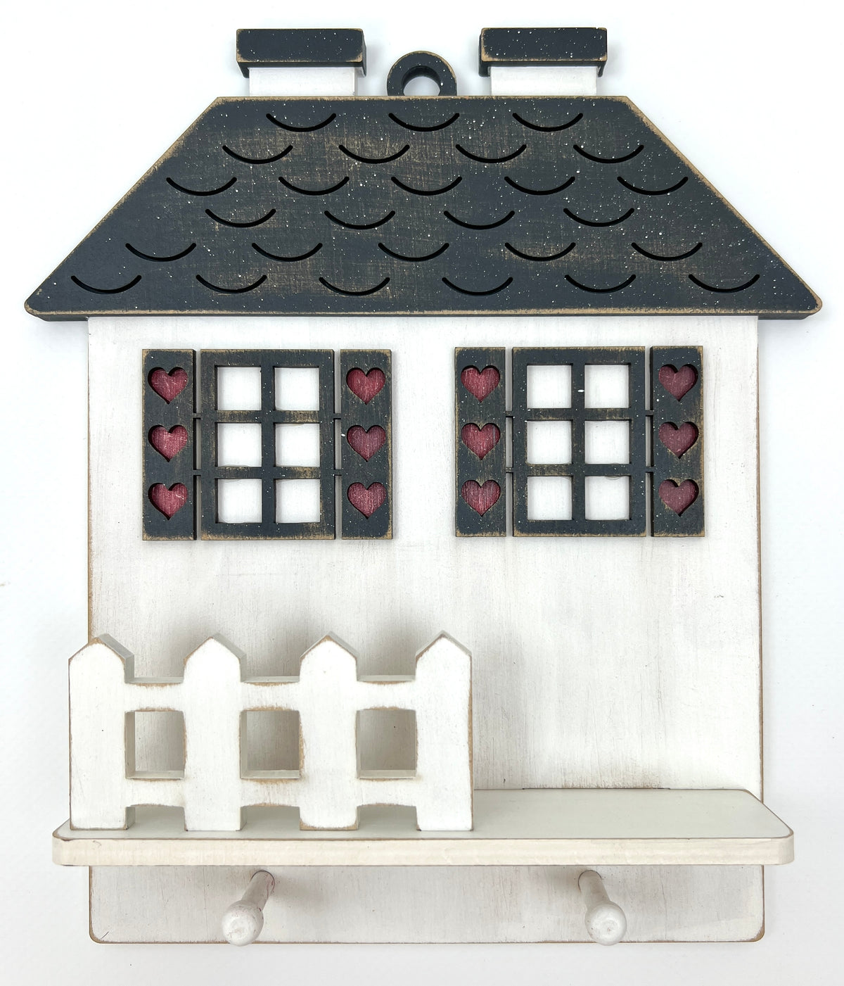 A decorative house-shaped wall shelf or ornament with a distressed white body, dark charcoal roof, two window panels with red heart cutouts, and a small white picket fence on the front.