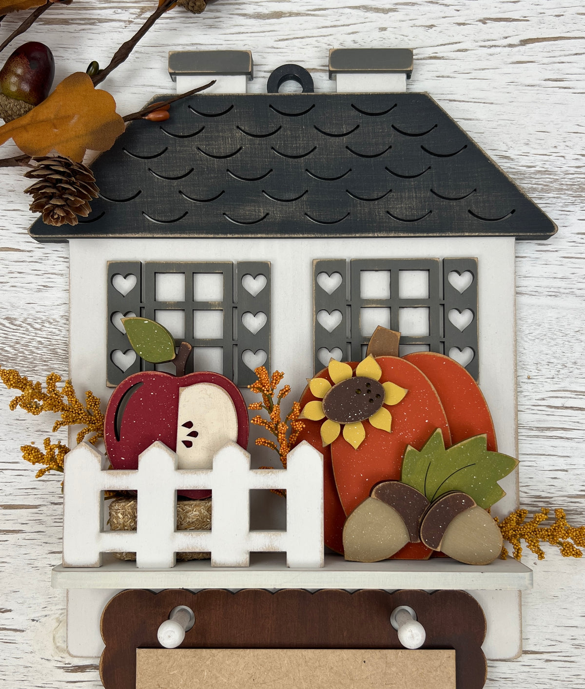A decorative autumn-themed wooden house ornament with a dark gray scalloped roof, white picket fence, apple, sunflower, pumpkin, acorns, and dried floral accents, displayed against a whitewashed wood background.