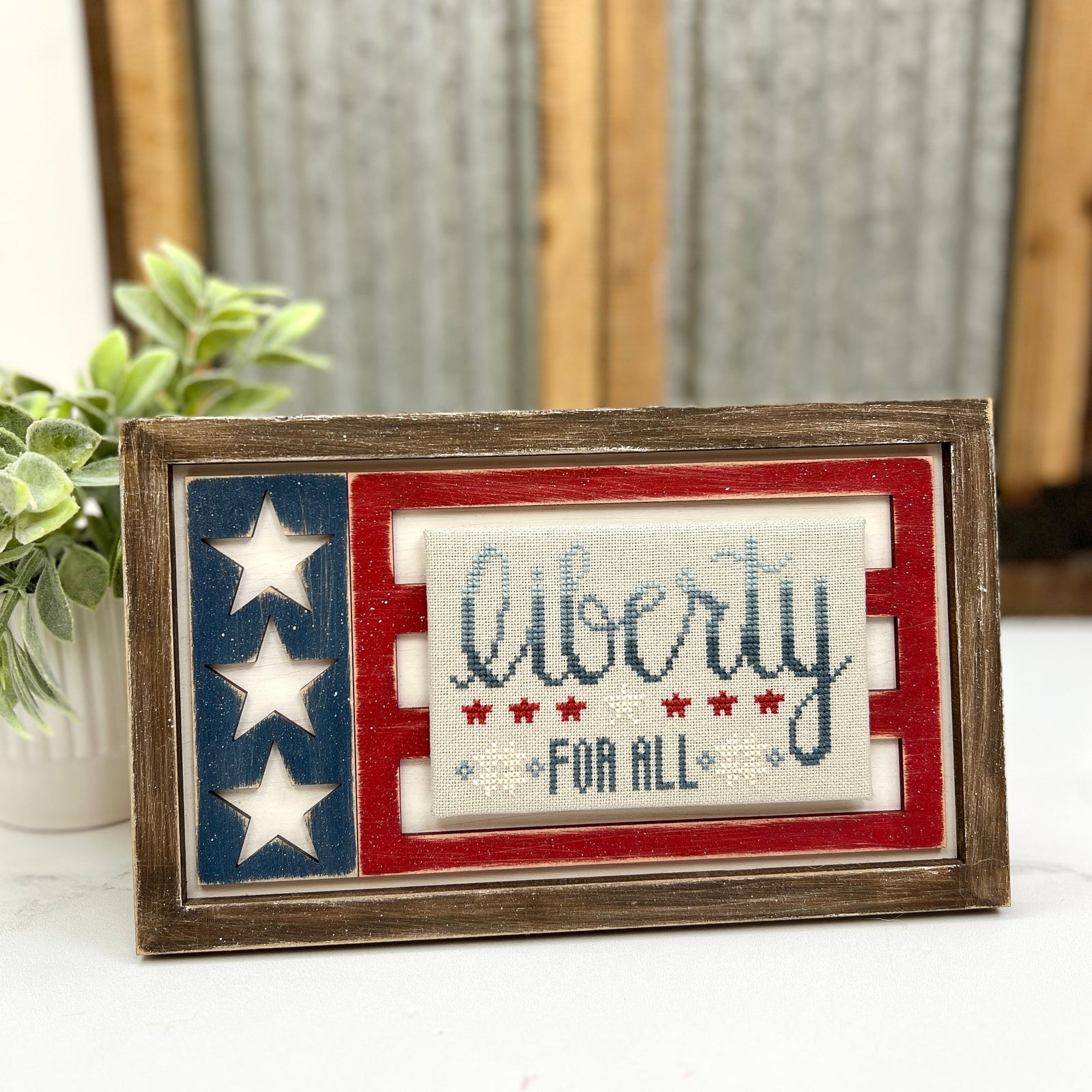 DIY wood cross stitch display frame painted in red, white, and blue with cutout stars on the left and a stitched piece in the center reading “liberty for all,” set on a rustic table with a plant in the background.