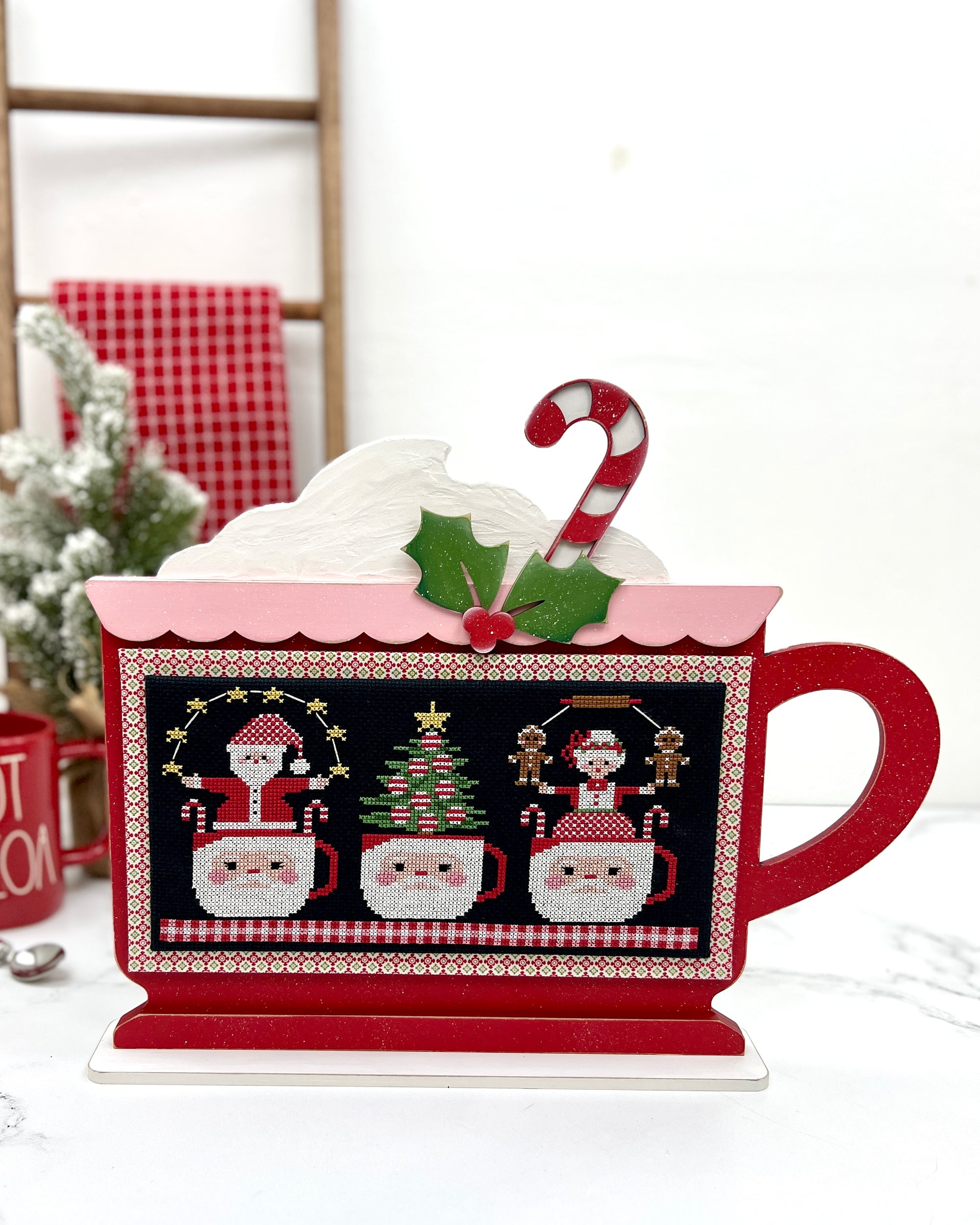 Cup O’ Christmas Cheer DIY wood cross stitch display shaped like a festive cocoa mug, shown with “Cups of Christmas Cheer” pattern by Stitching with the Housewives.