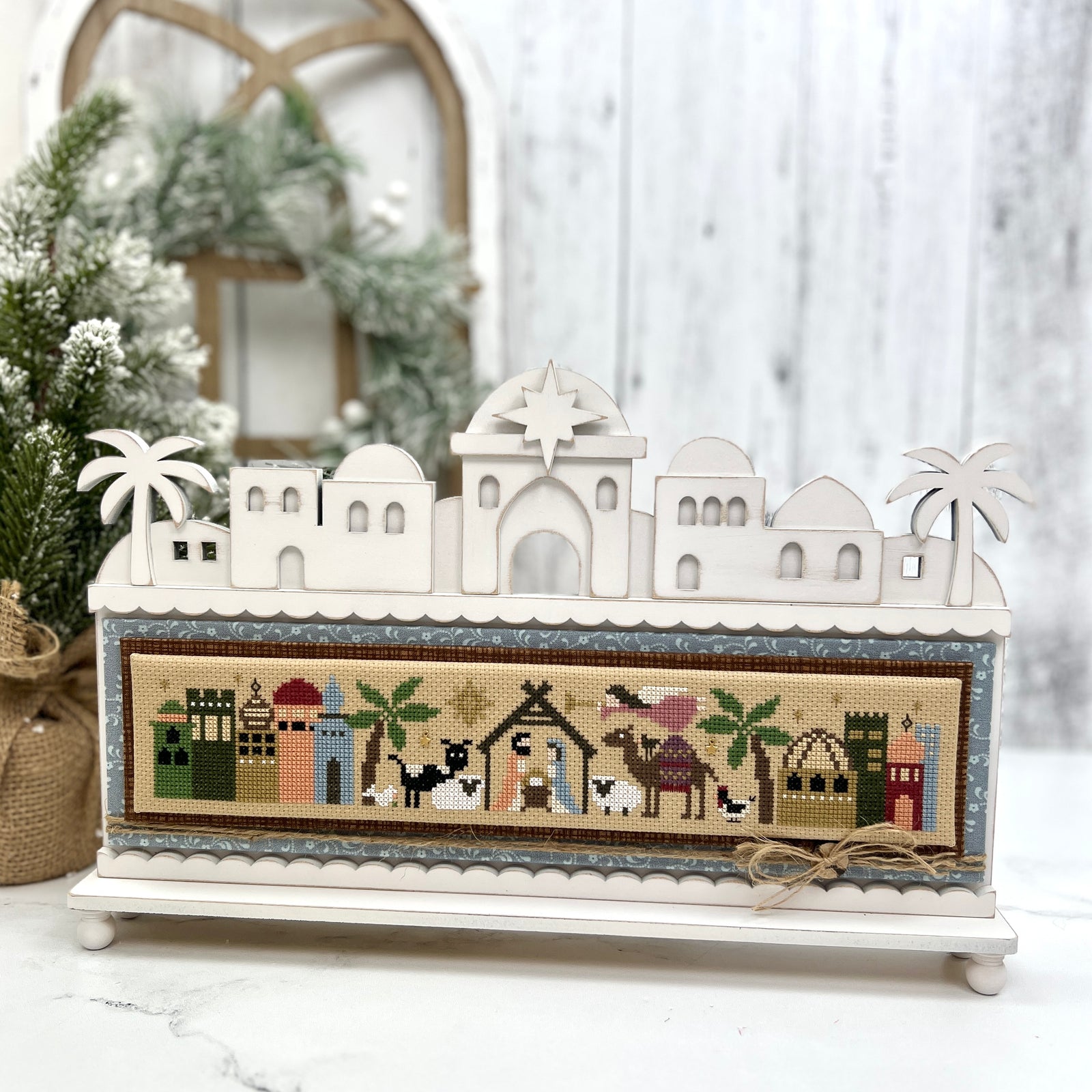 DIY Bethlehem nativity skyline wood display backer for cross stitch projects, with layered rooftops, arched windows, palm trees, and Star of Bethlehem. Cross stitch called Nativity Tiny Town by Heart in Hand