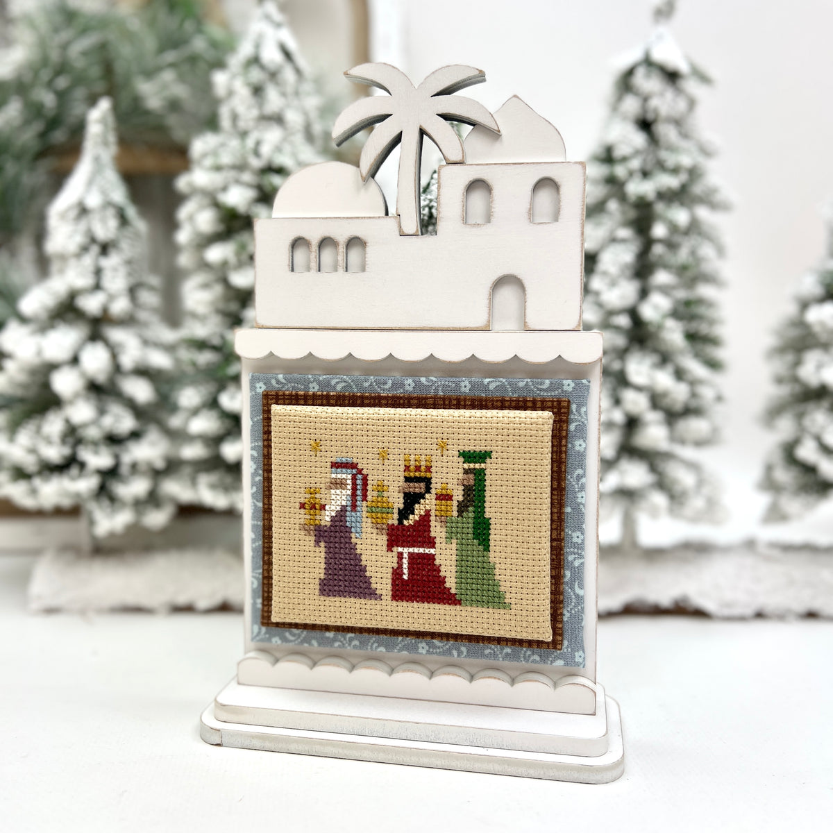 Decorative scene with a white building, palm tree, and nativity scene on a stand against a snowy background.