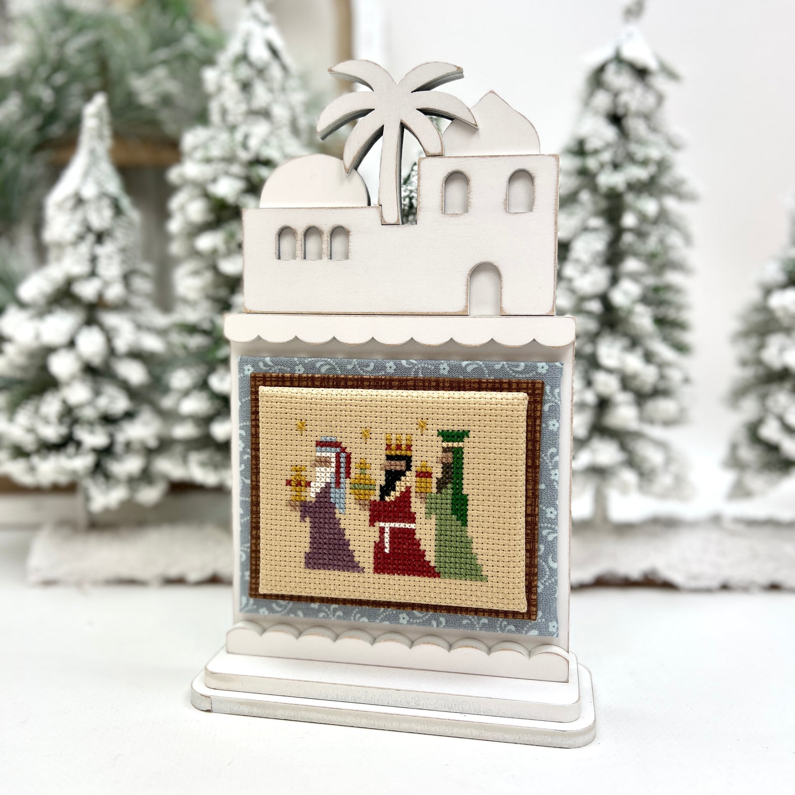 Decorative scene with a white building, palm tree, and nativity scene on a stand against a snowy background.