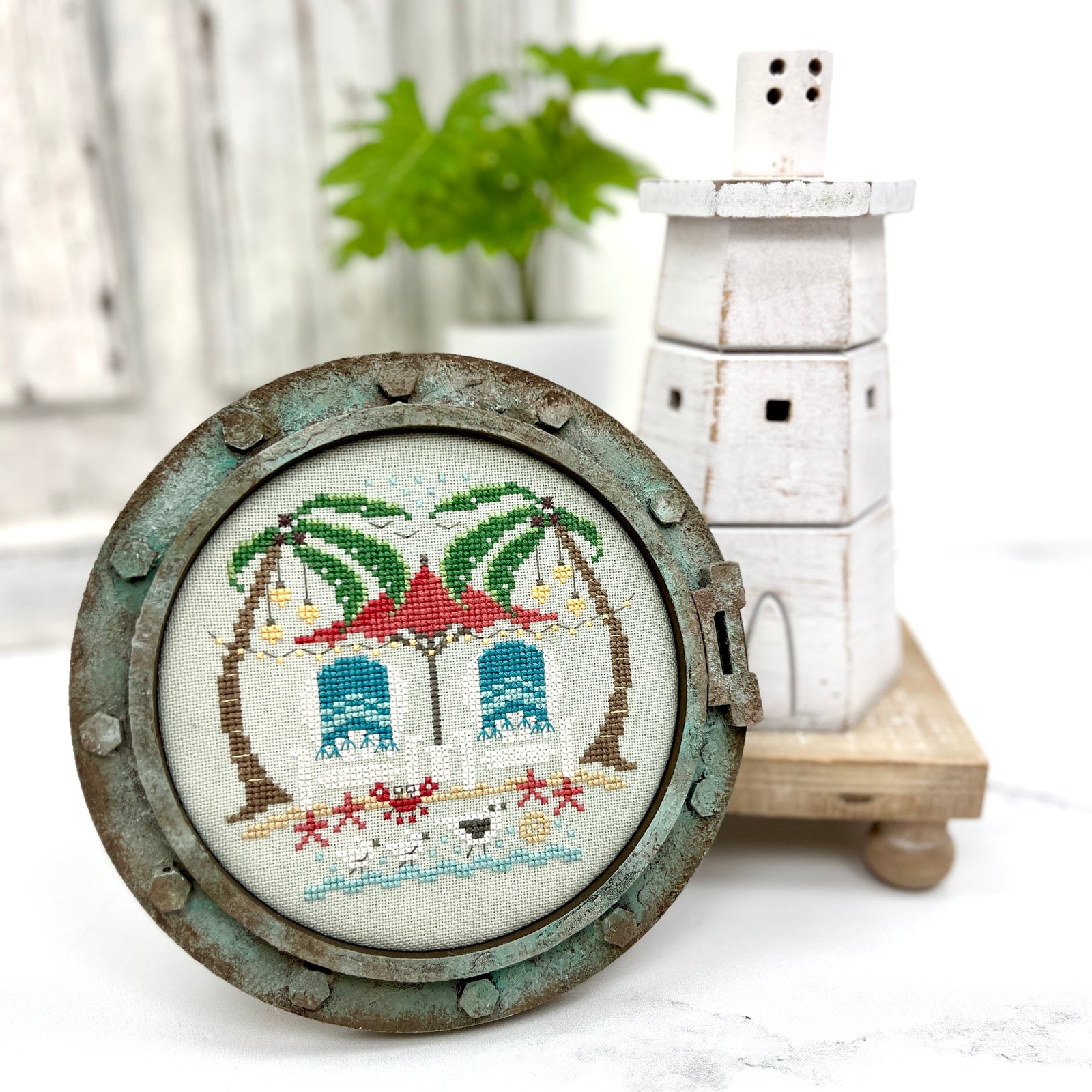 Decorative cross-stitched scene with palm trees and buildings in a round metal frame. Cross stitch is called toes in the sand by Hands on Design.