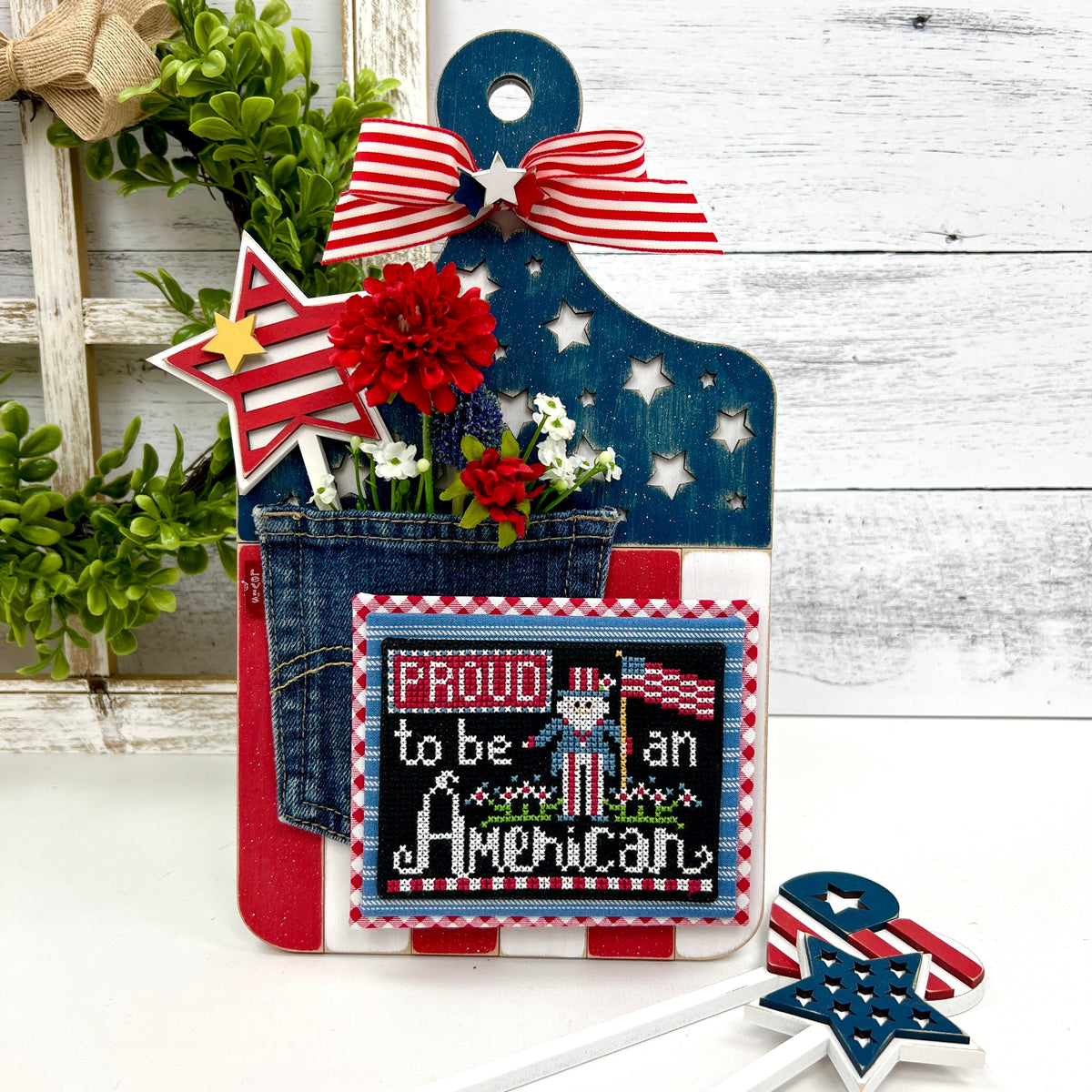 Patriotic themed wood paddle board cross stitch display painted red, white, and blue. Accents of stars, a jean pocket and flowers. Proud to be an American cross stitch by Stitching with the Housewives.