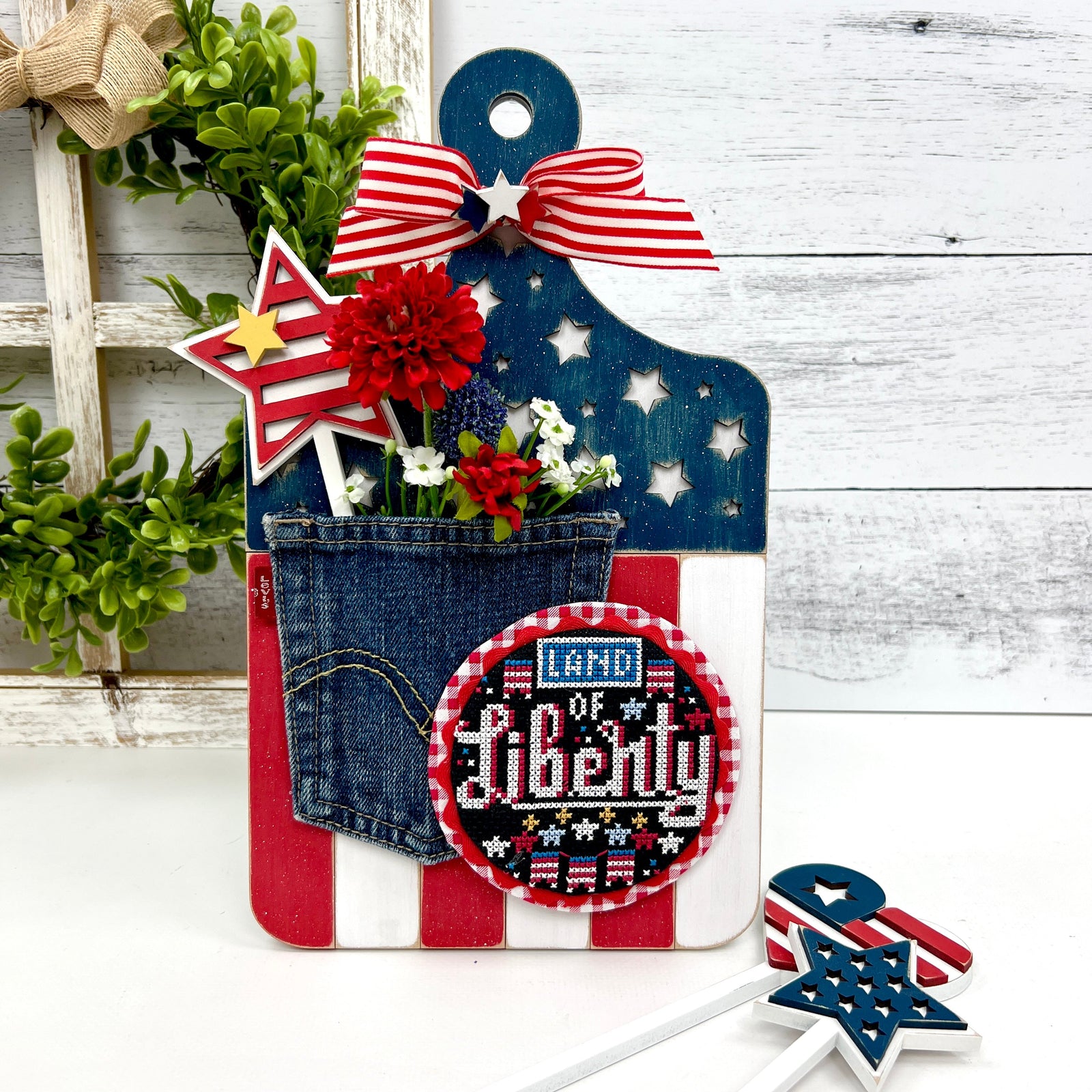 Patriotic themed wood paddle board cross stitch display painted red, white, and blue. Accents of stars, a jean pocket and flowers. Liberty cross stitch by Stitching with the Housewives.