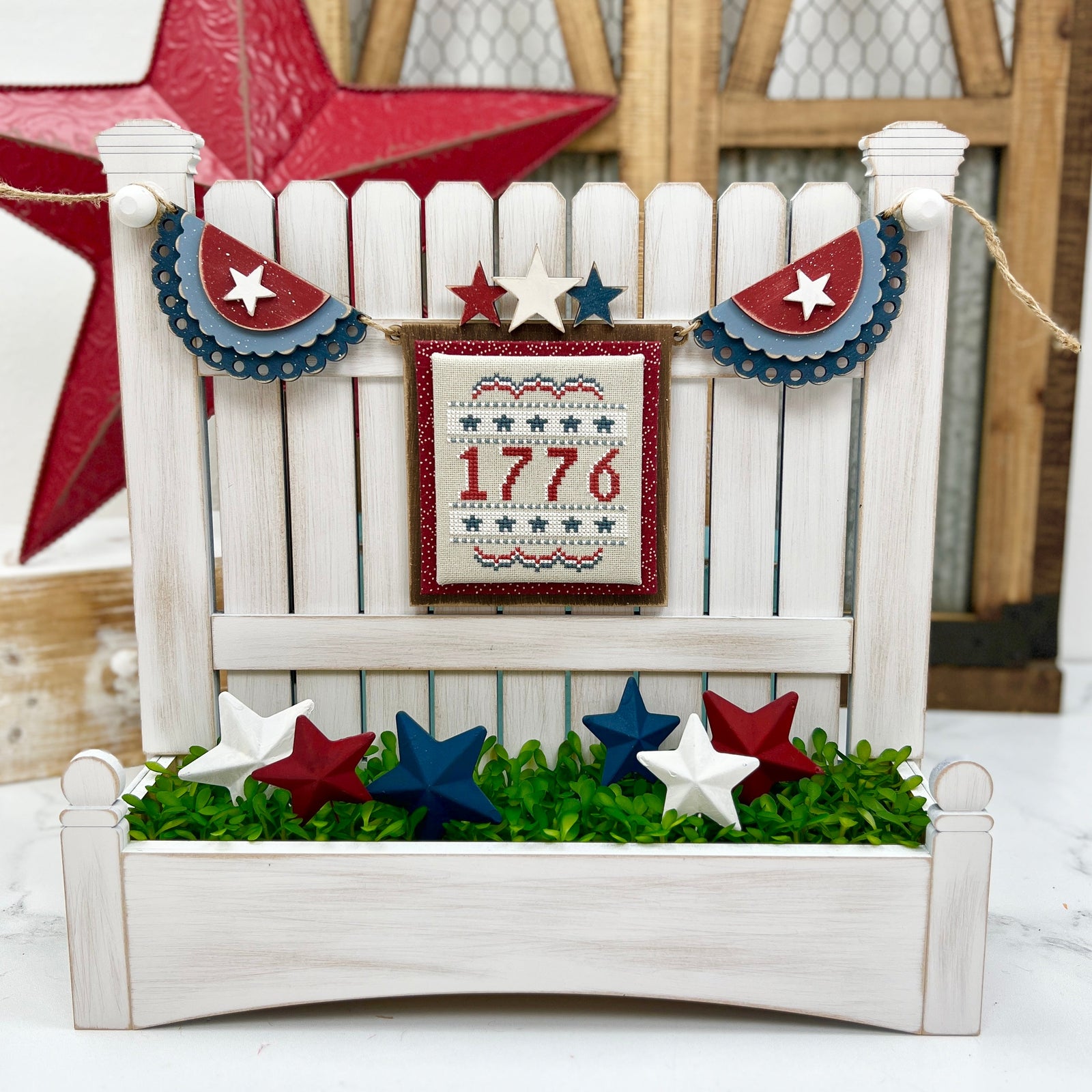 DIY patriotic garland with layered red, white, and blue wooden bunting and stars, strung across a white picket fence cross stitch display with 1776 stitching and 3D stars in a faux grass planter.