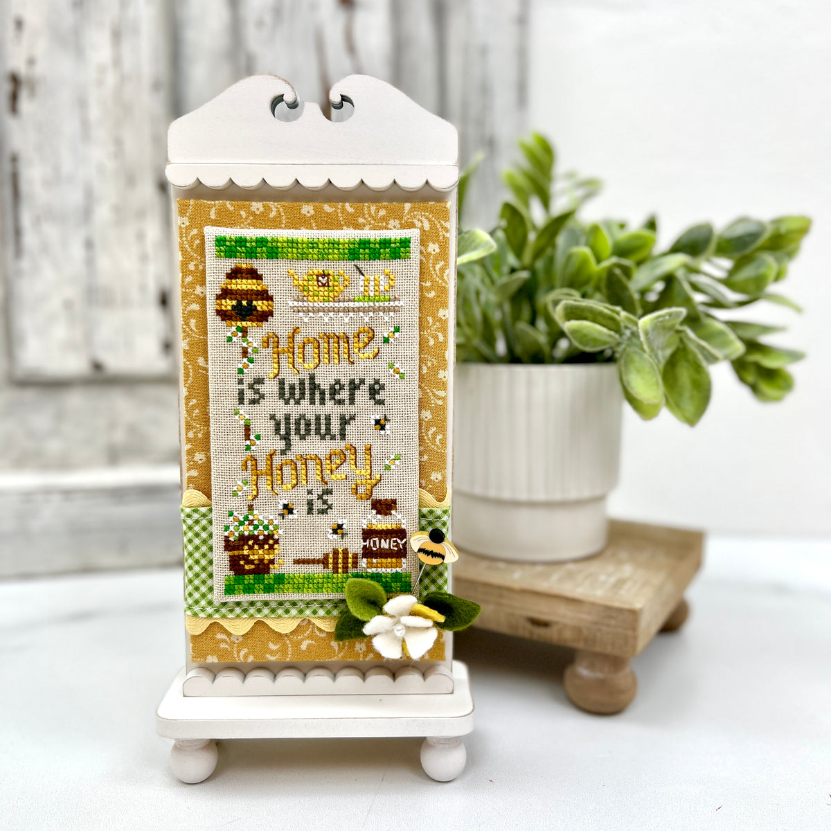 Tall cabinet-style wood cross stitch display frame with scalloped crest top and pedestal base, shown holding a honeybee themed cross stitch.