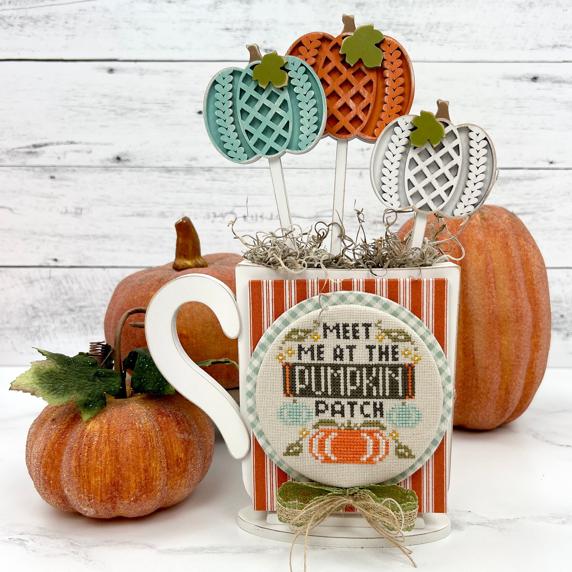 Wood pumpkins in a mug with a finished cross stitch that says meet me at the pumpkin patch by Stitching with the Housewives.