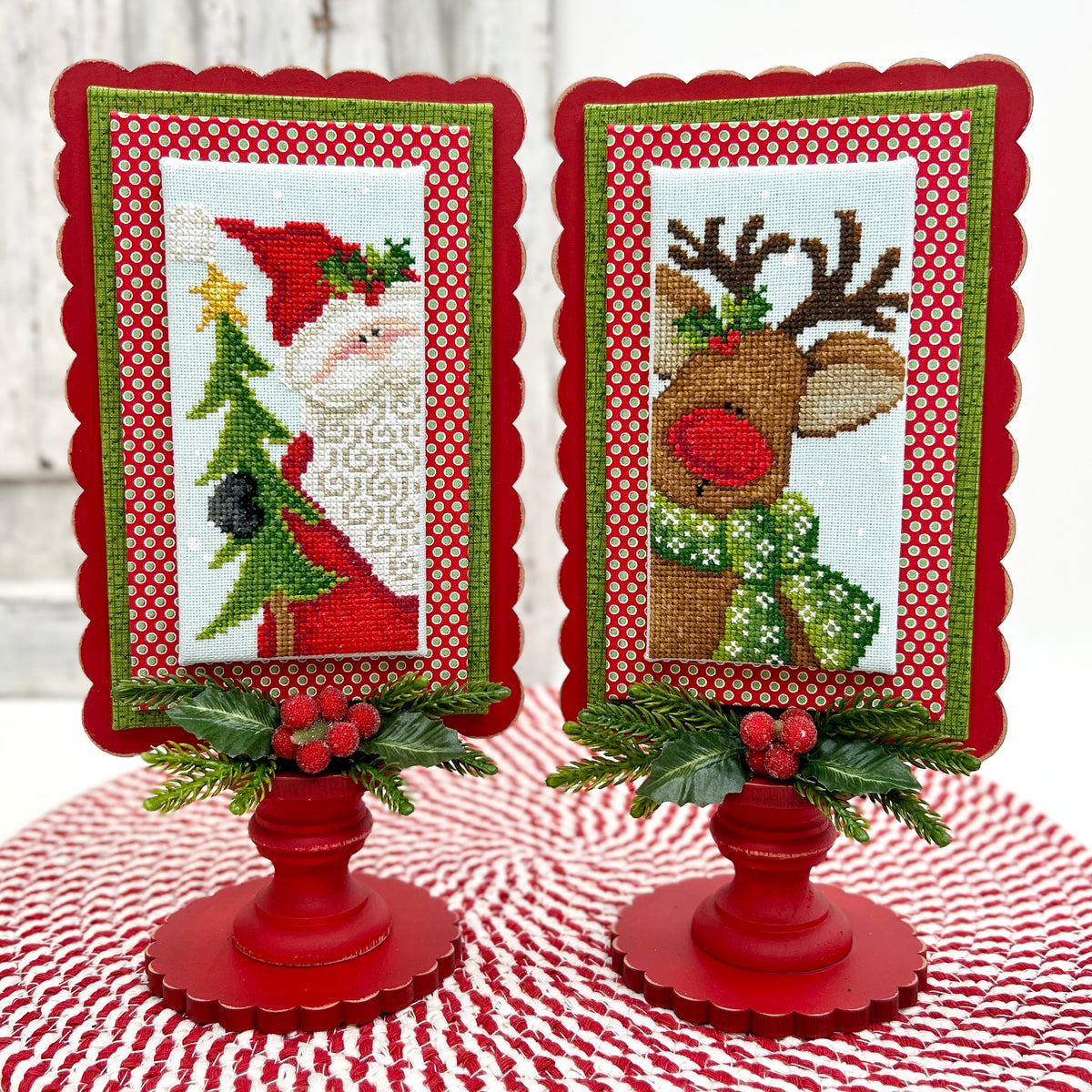 Two Christmas-themed cross-stitch frames on red stands with a white background.