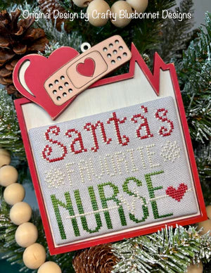 Close-up of the Santa’s Favorite Nurse wood ornament display featuring a red frame, heart accent, and bandage detail, holding a festive nurse-themed cross stitch design among holiday greenery.