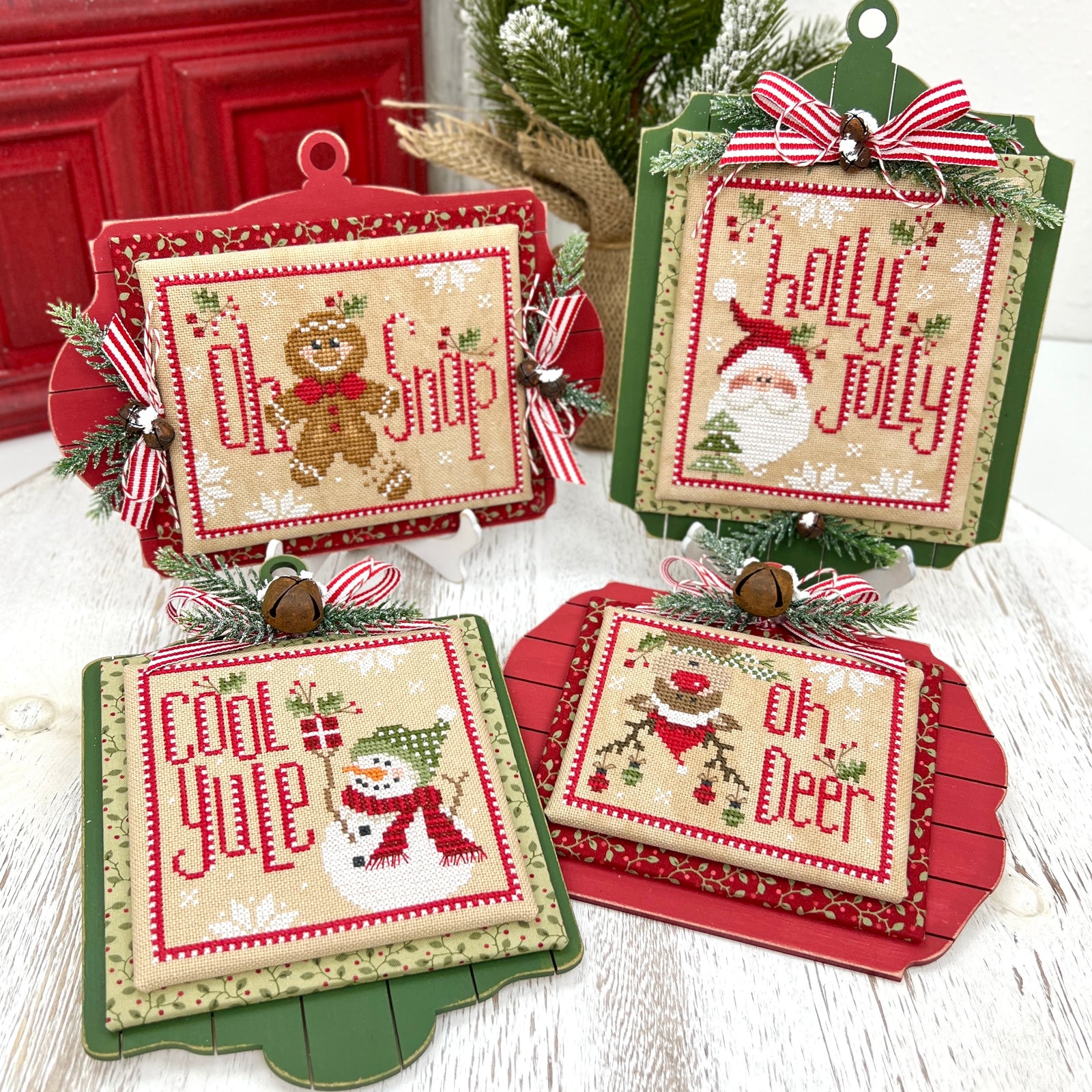 Four Christmas-themed cross-stitch ornaments arranged on a white wooden surface. The cards feature festive designs including a gingerbread man, Santa Claus, a snowman, and holiday text, with red and green frames, striped bows, pine branches, and small bells.
