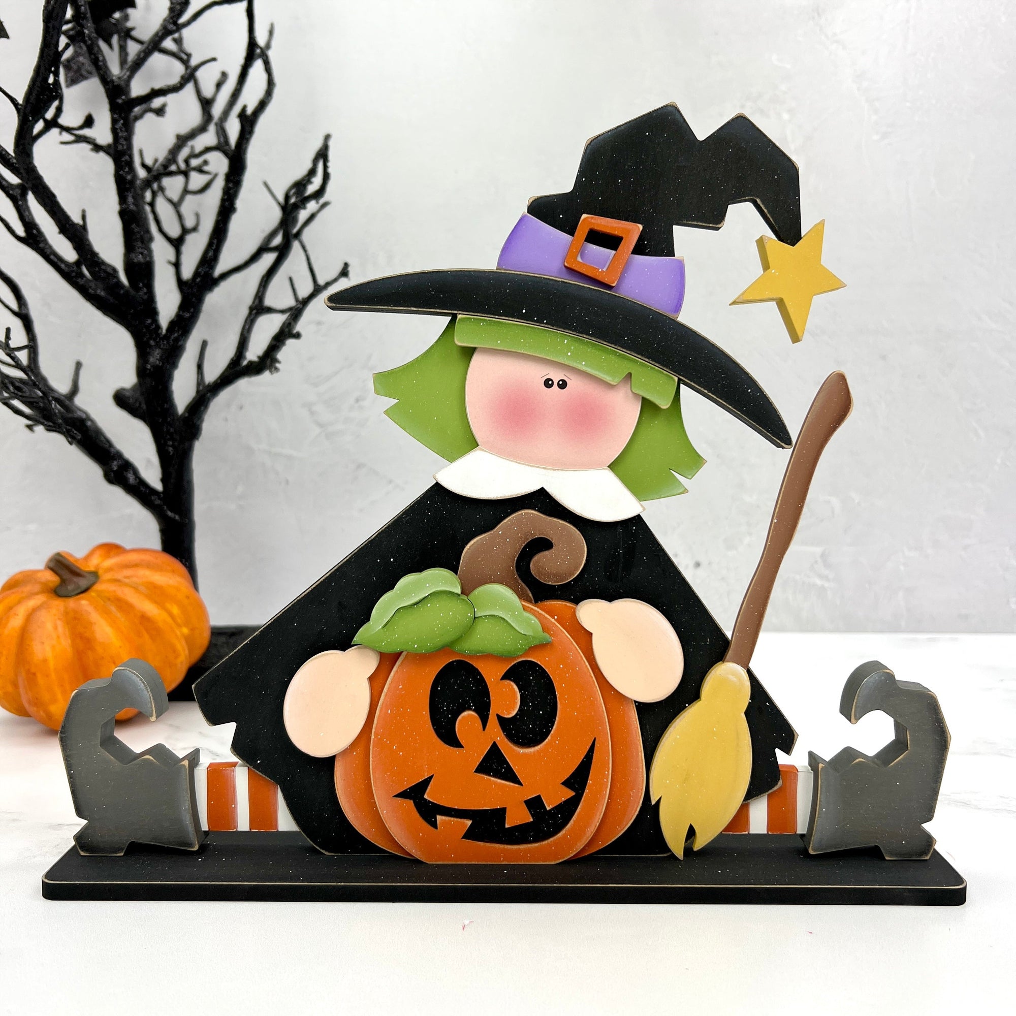 Sitting witch holding a funny face pumpkin and broomstick wood decor craft kit. 