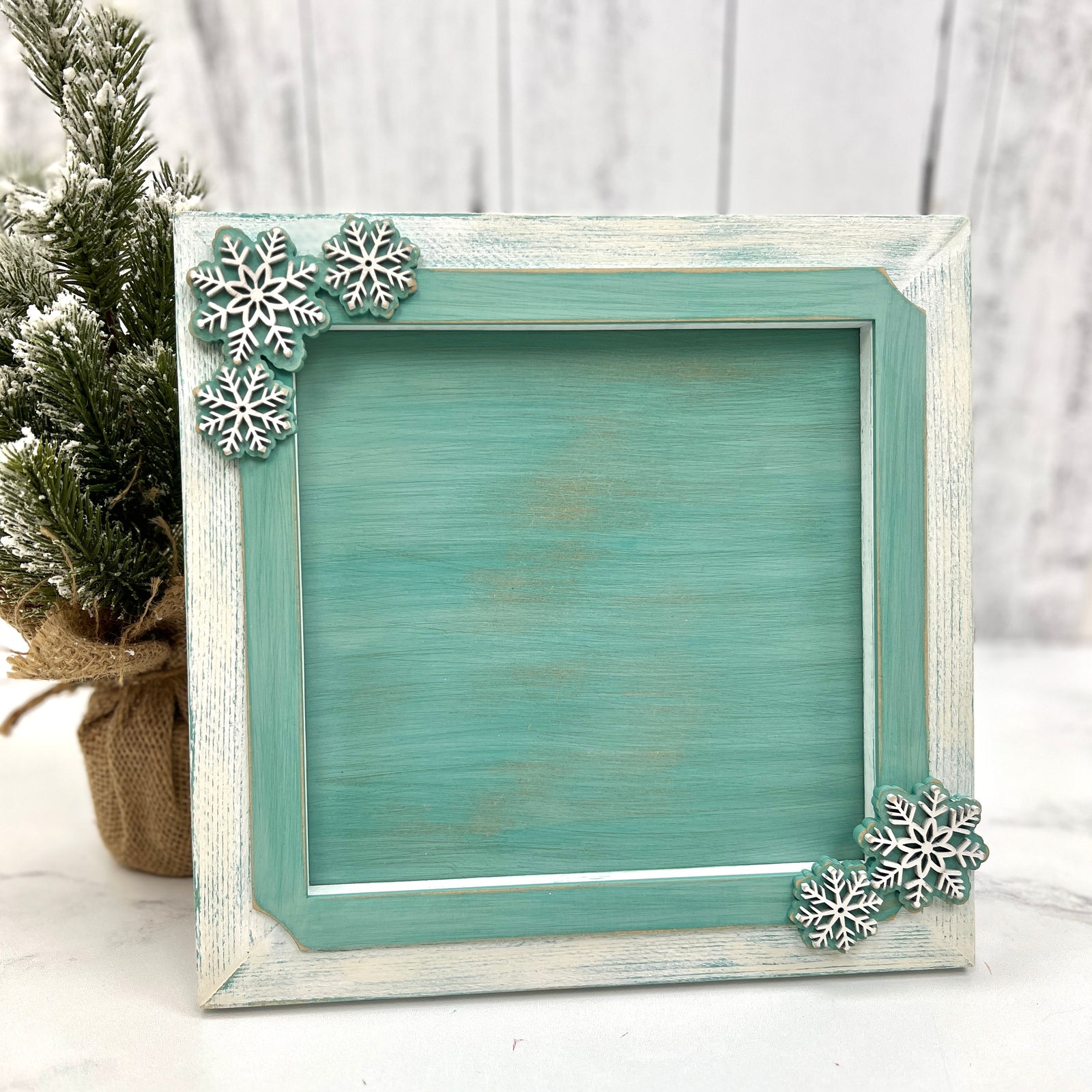 Rustic winter-themed wood frame painted in distressed aqua and white, accented with decorative snowflakes in the corners, designed to hold a finished cross stitch piece.