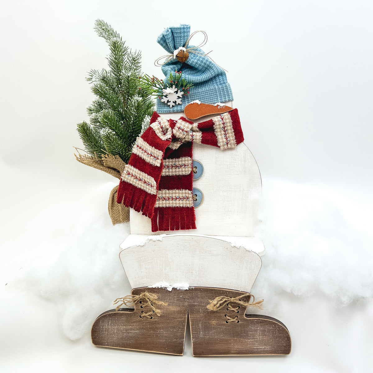 Snowman shelf sitter made of painted wood, wearing a blue fabric hat and red scarf, accented with greenery and faux snow, designed as a winter farmhouse display for shelves or tabletops.