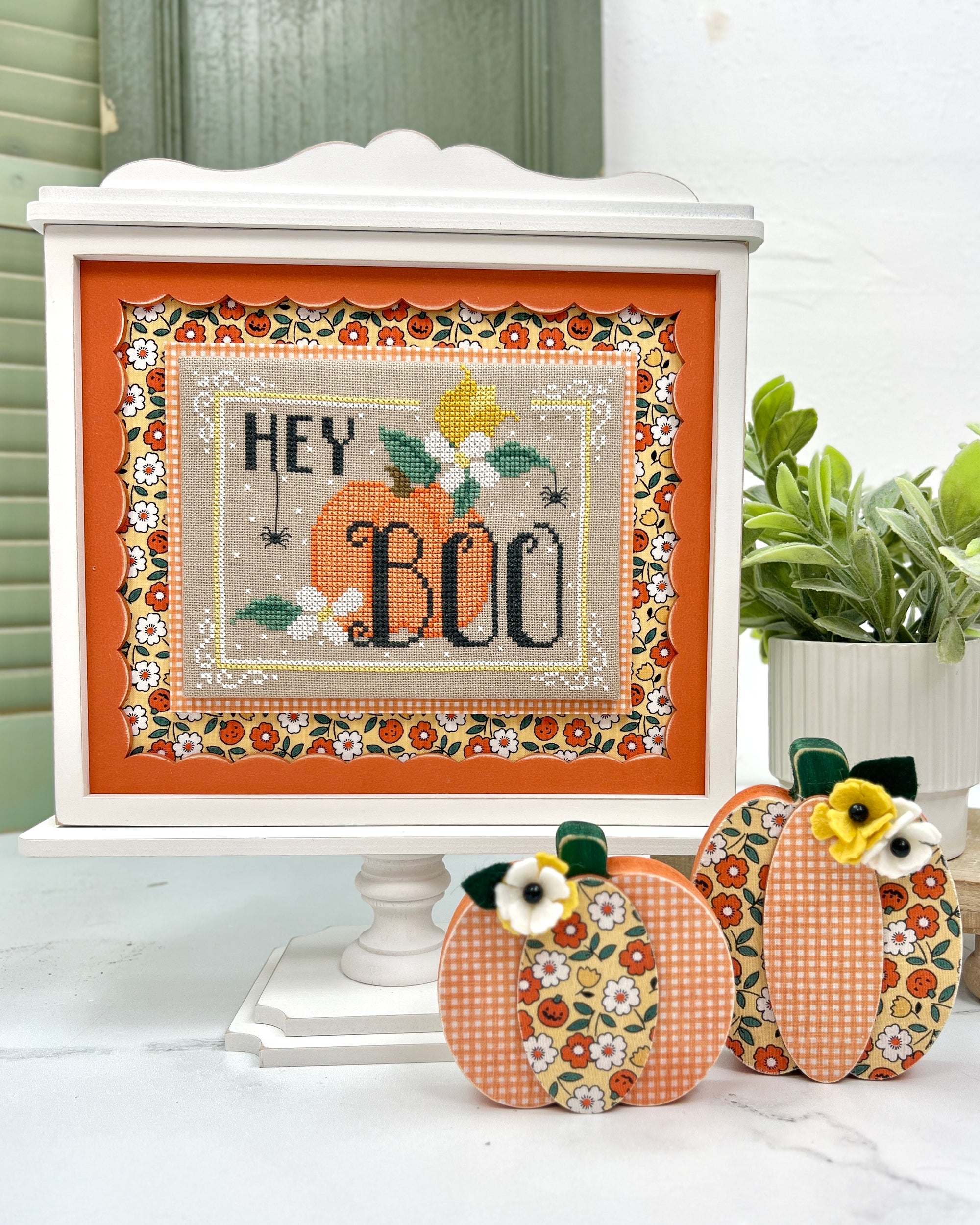 Unfinished wood pedestal cross stitch display frame with scalloped edge insert and wide recessed border. Freestanding design with decorative pedestal base, ready to paint any color to match seasonal or themed stitching projects.