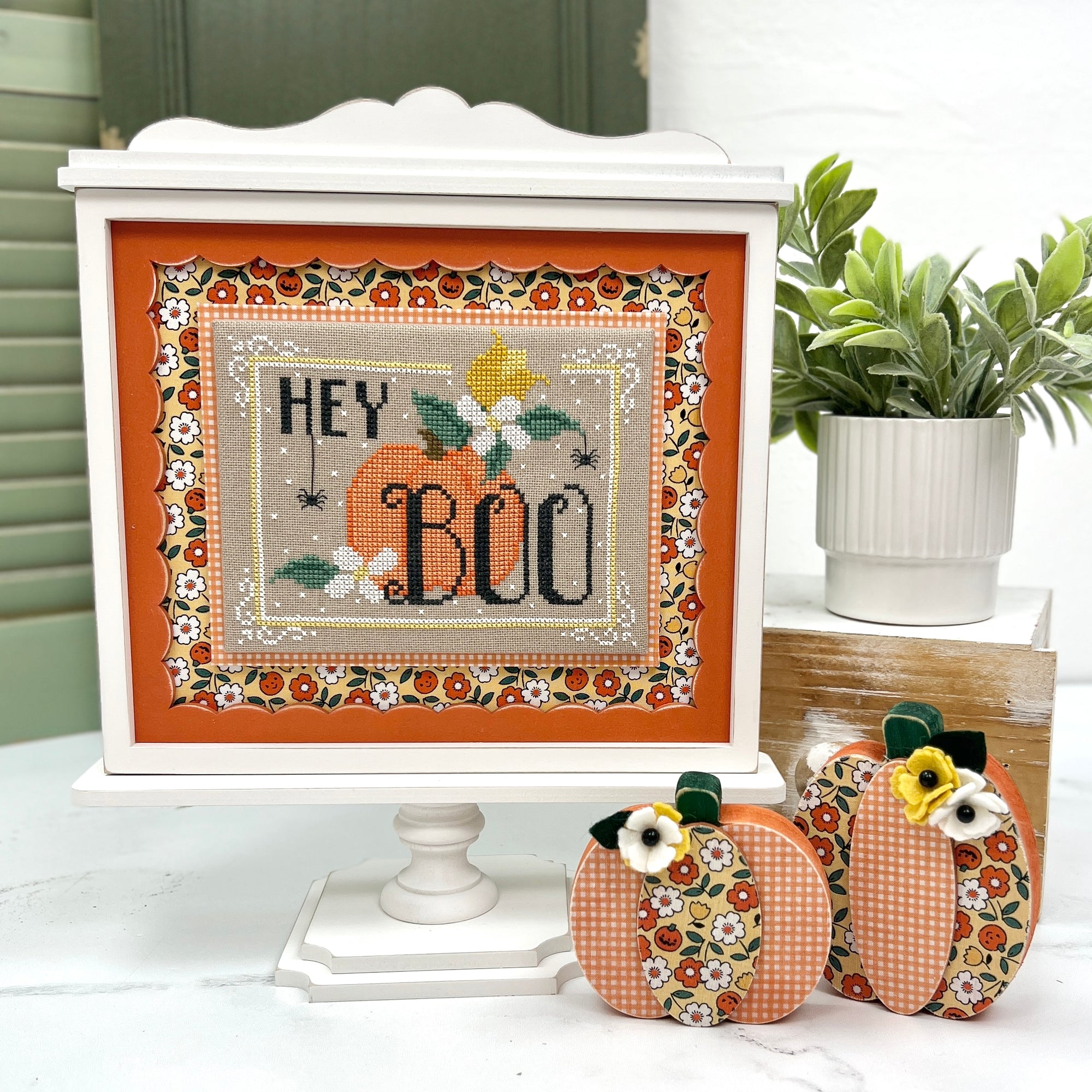Decorative cross-stitch piece with pumpkins and 'Hey Boo' text on a white pedestal, surrounded by small decorative pumpkins and a plant.