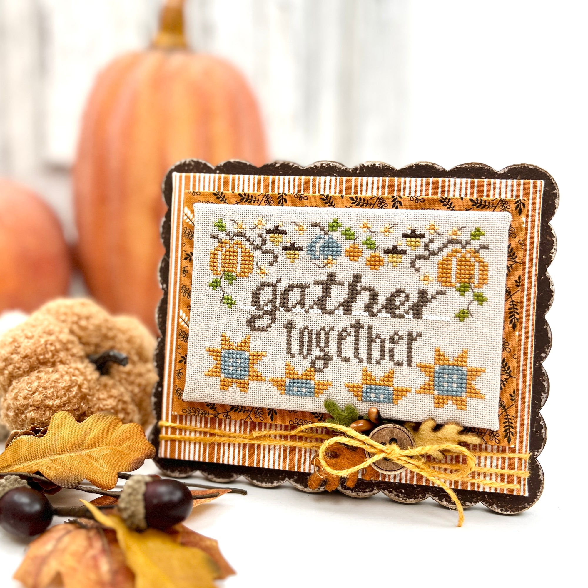 Cross-stitched piece with 'gather together' text, surrounded by autumn decorations.